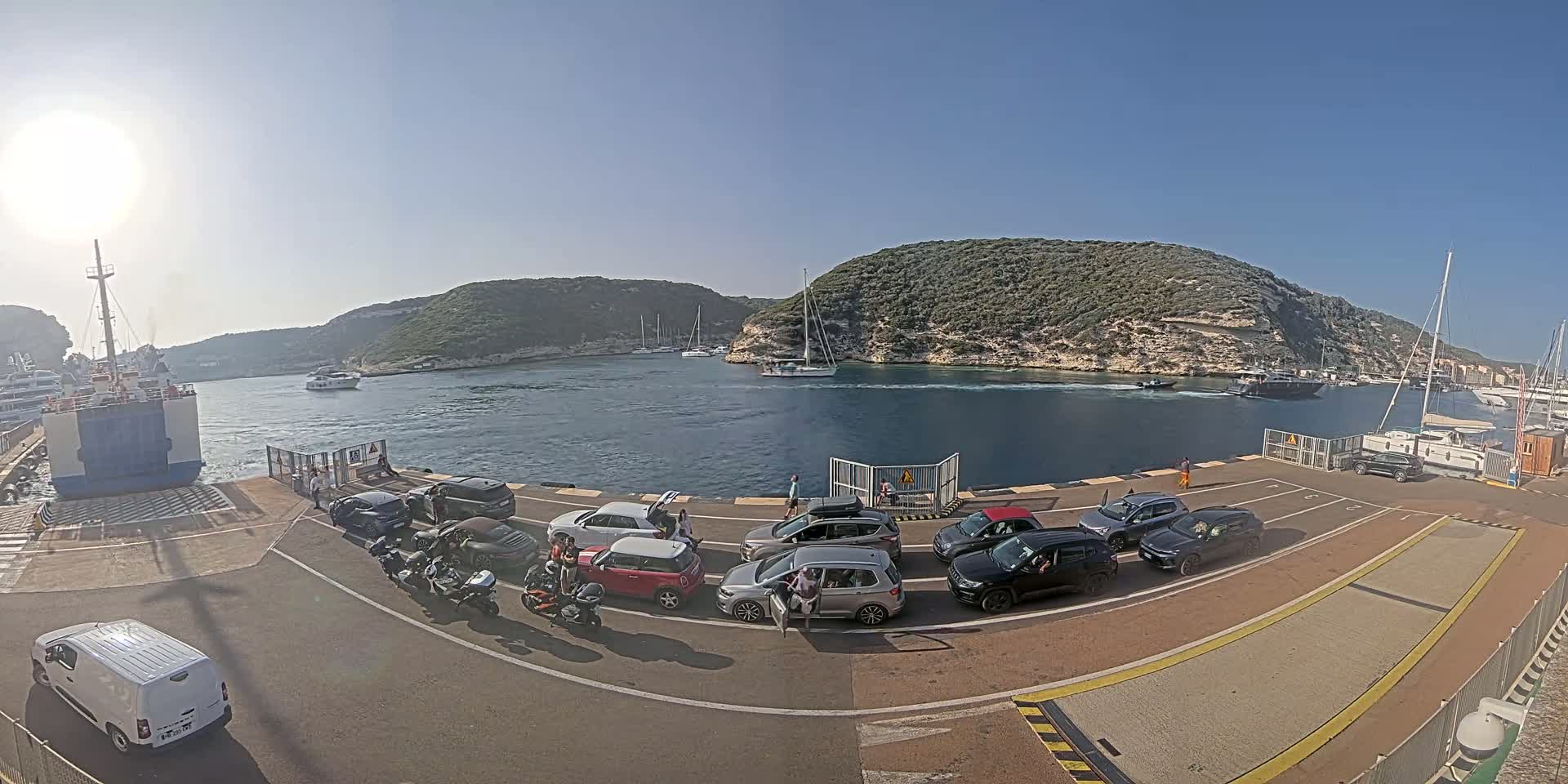 A ferry is docked at a port on a sunny day, with several cars and motorcycles waiting to board, surrounded by calm water and rocky hills.