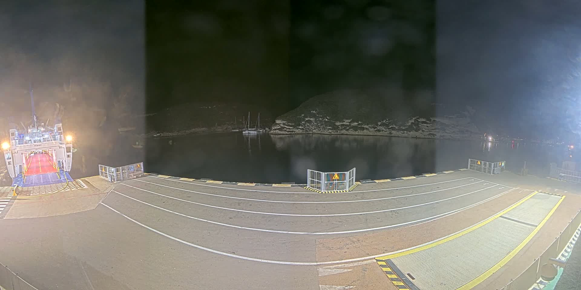A ferry is docked at a port on a sunny day, with several cars and motorcycles waiting to board, surrounded by calm water and rocky hills.