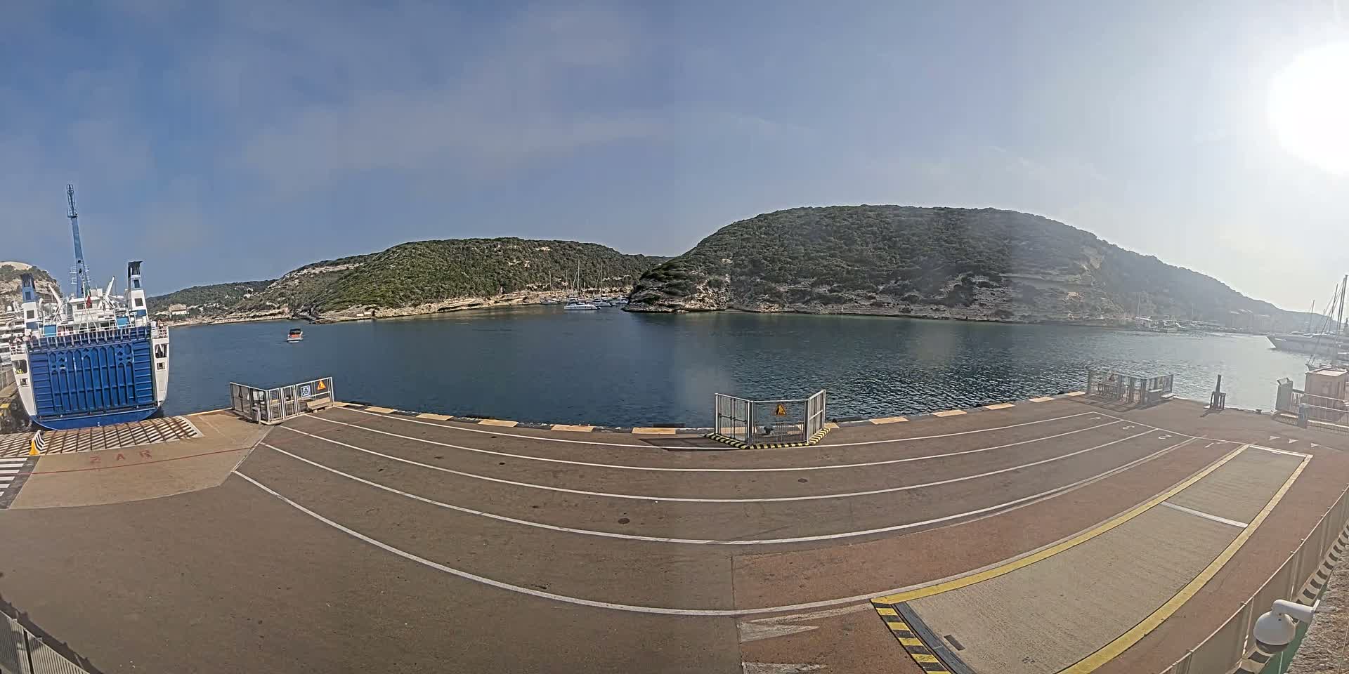 A ferry is docked at a port, with calm water and hills covered in green vegetation visible in the background under a clear, sunny sky.