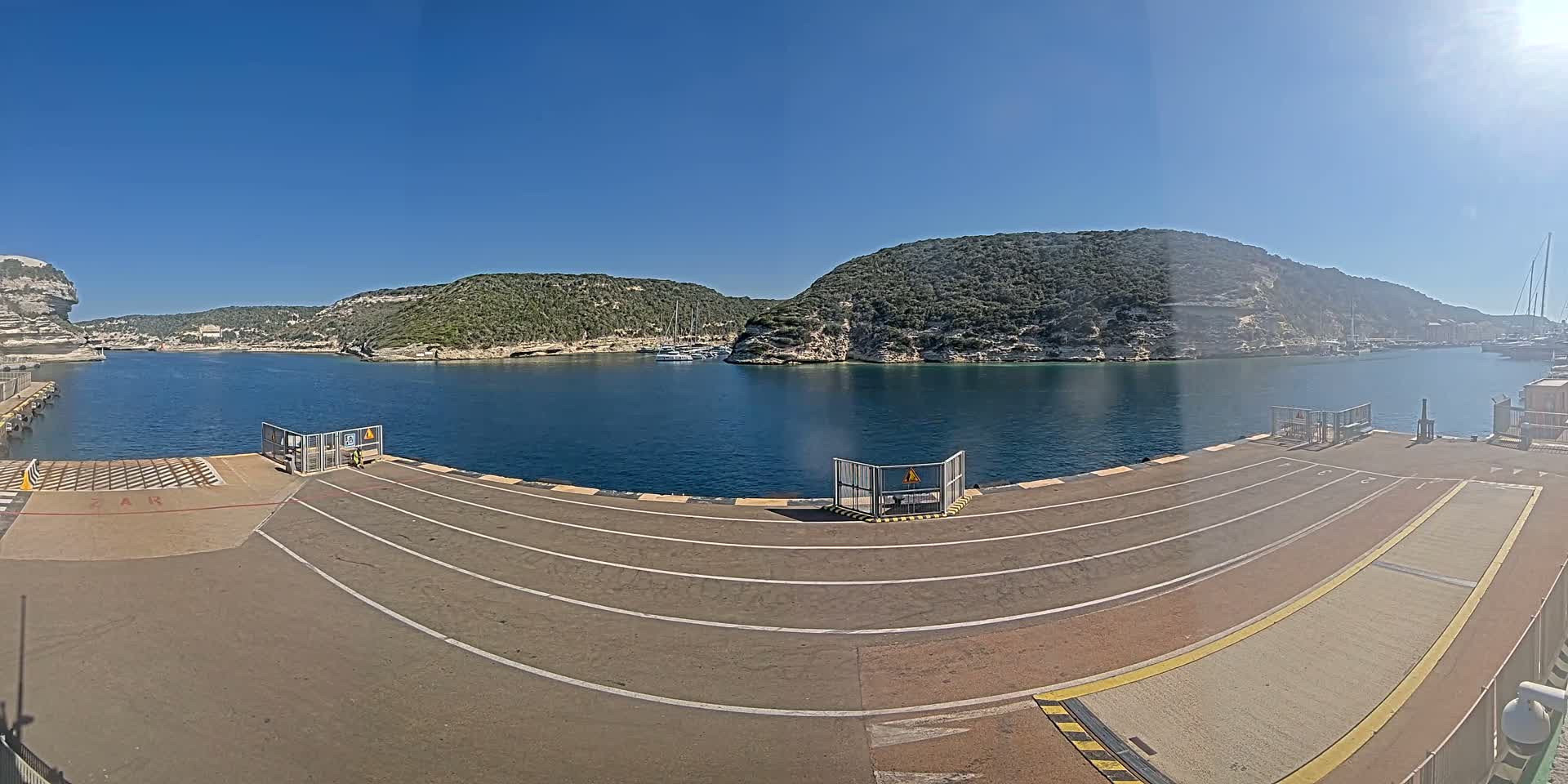 A ferry dock with marked lanes overlooks a calm bay surrounded by green hills and several moored boats under a clear blue sky.