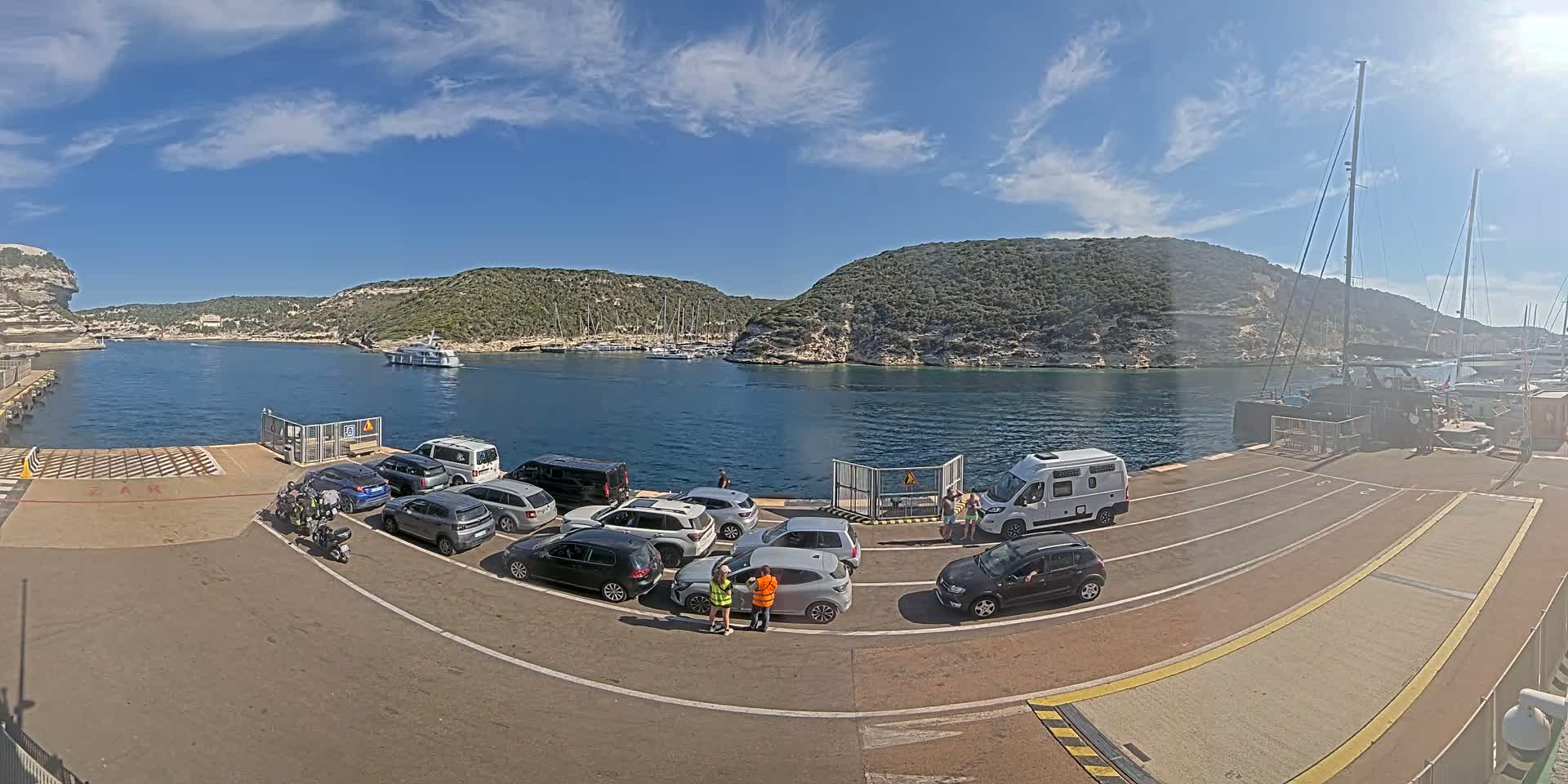 A ferry terminal with several cars and a camper van waiting to board, situated on a calm body of water under a sunny sky with scattered clouds, surrounded by green hills.