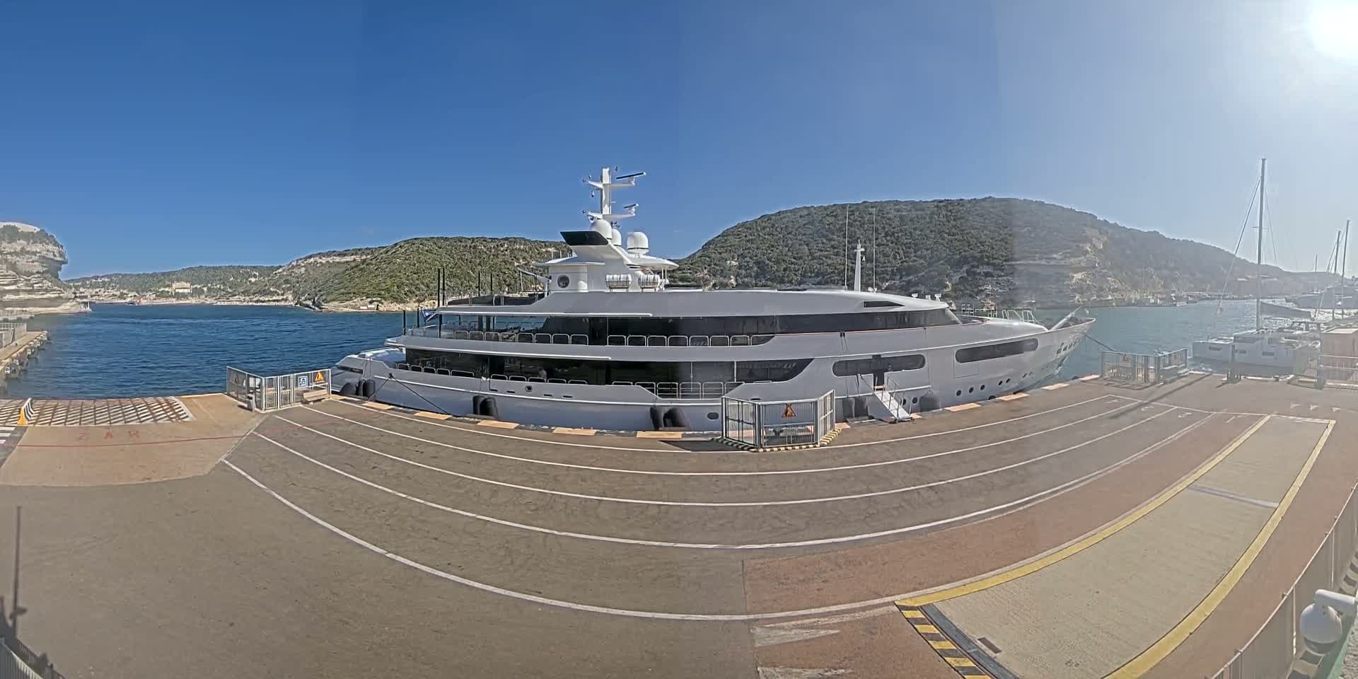A large white yacht is docked at a port on a sunny day, with calm water and hills in the background.
