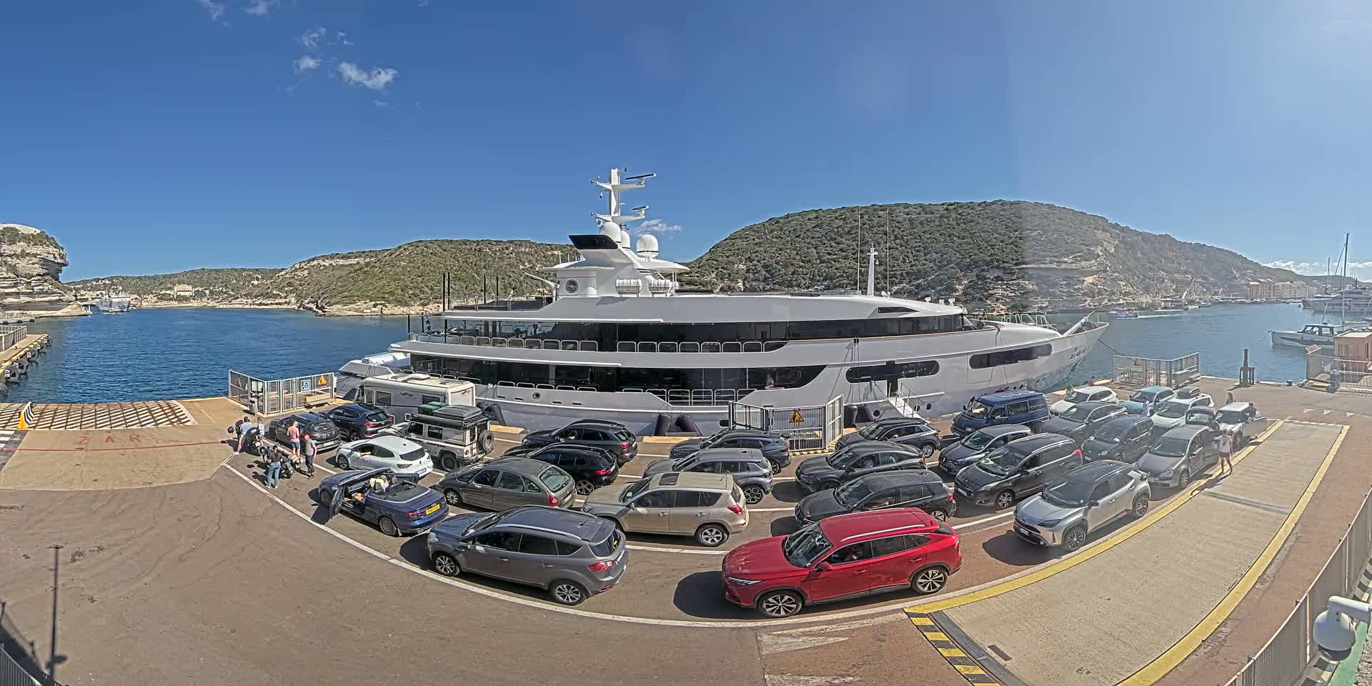 A large yacht is docked at a port on a sunny day, with numerous cars lined up on the dock awaiting embarkation or disembarkation.