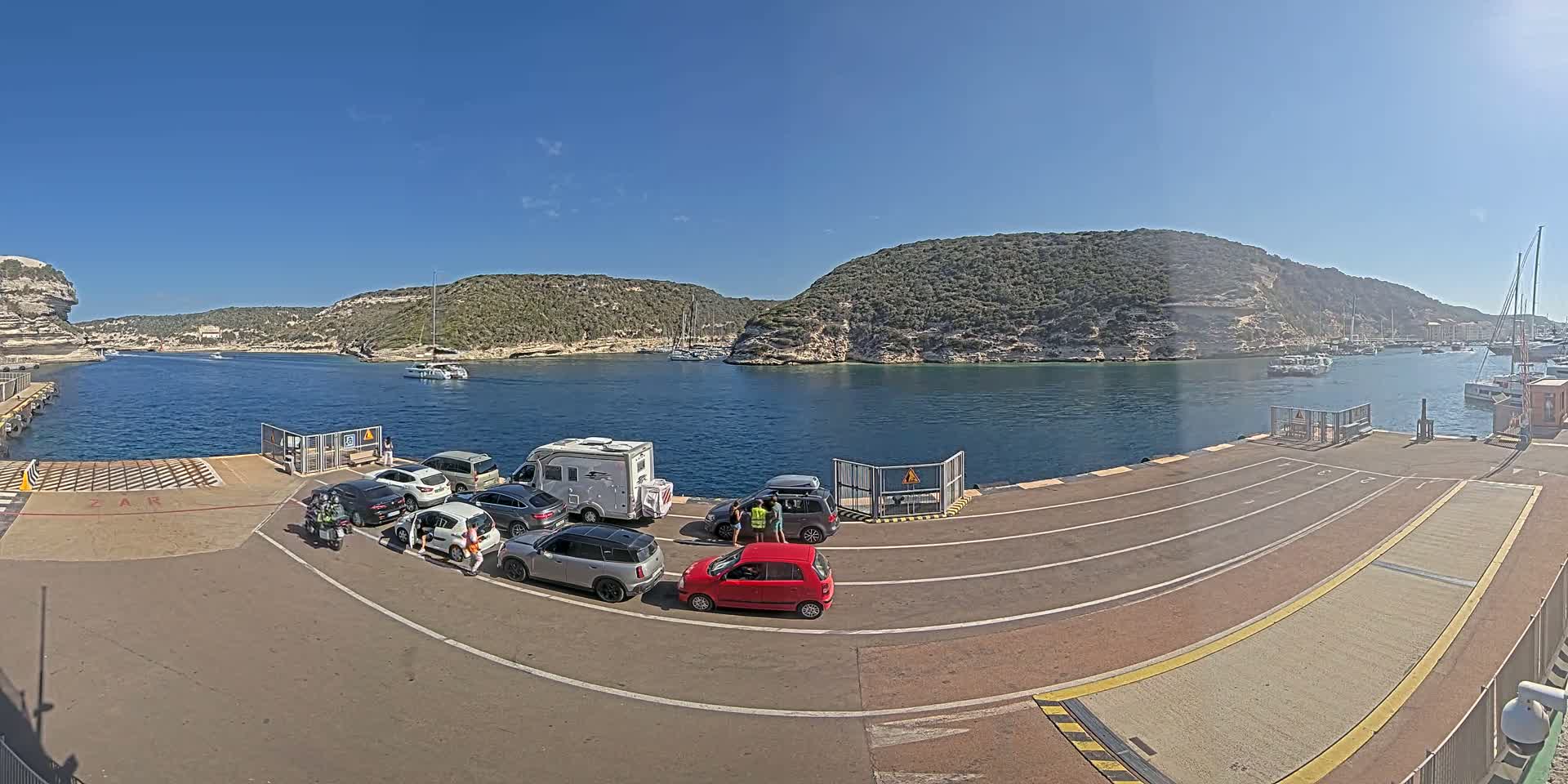 Several cars and a campervan are parked on a ferry dock overlooking a calm bay with boats and rocky hills under a clear blue sky.