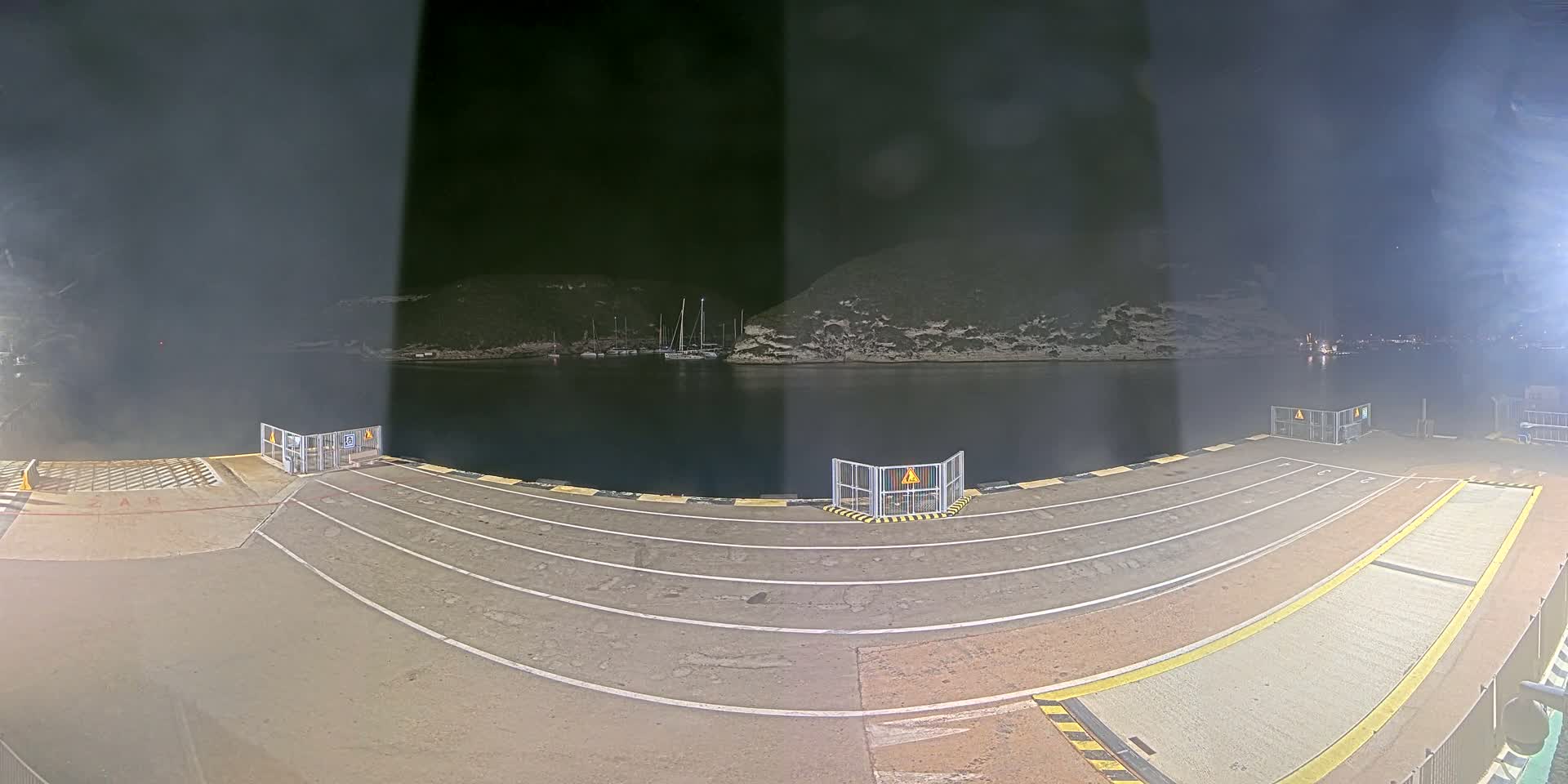 Several cars and a campervan are parked on a ferry dock overlooking a calm bay with boats and rocky hills under a clear blue sky.