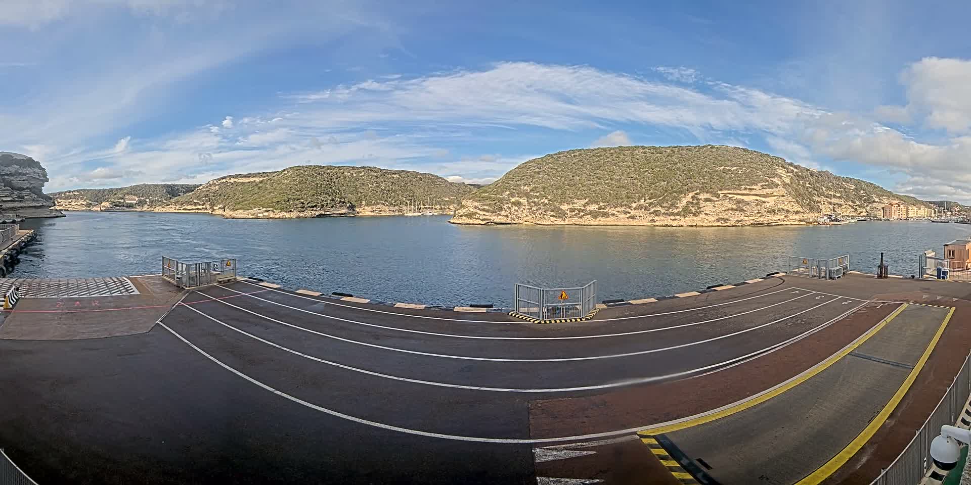 Several cars and a campervan are parked on a ferry dock overlooking a calm bay with boats and rocky hills under a clear blue sky.