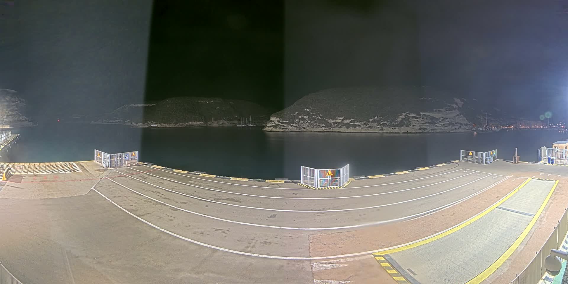 Several cars and a campervan are parked on a ferry dock overlooking a calm bay with boats and rocky hills under a clear blue sky.