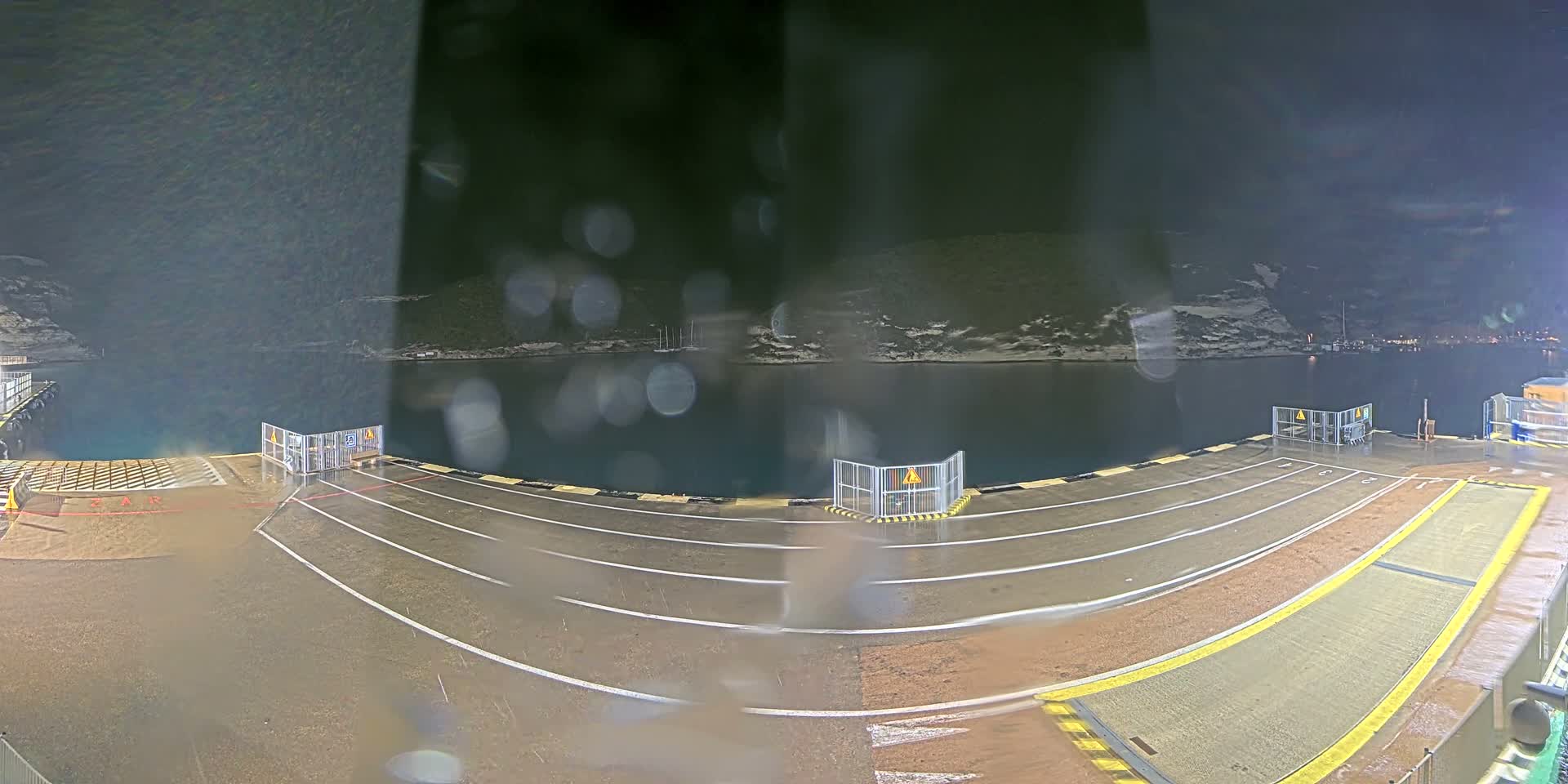 A wide-angle view captures a wet, empty ferry dock with lane markings and safety barriers extending towards dark water and distant, dimly lit landforms on a rainy night.
