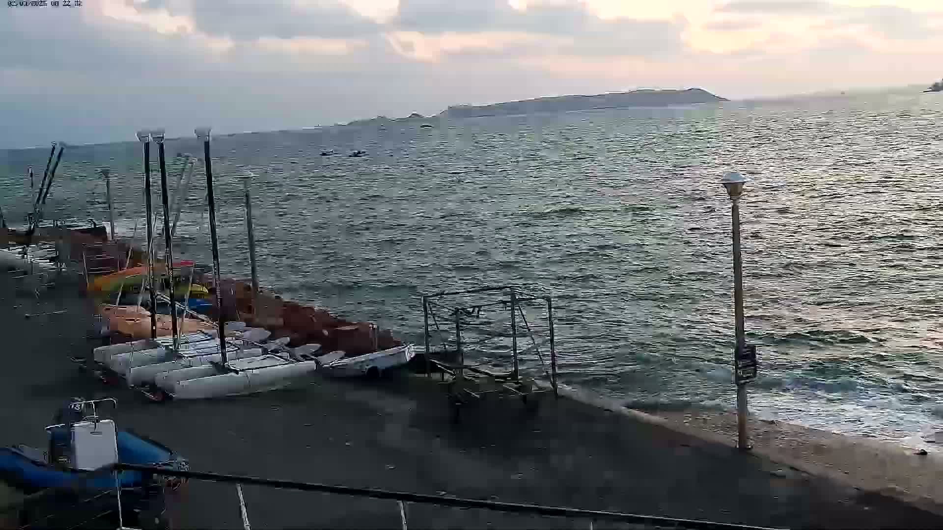 Several sailboats are docked along a shoreline with slightly choppy water under a cloudy sky.