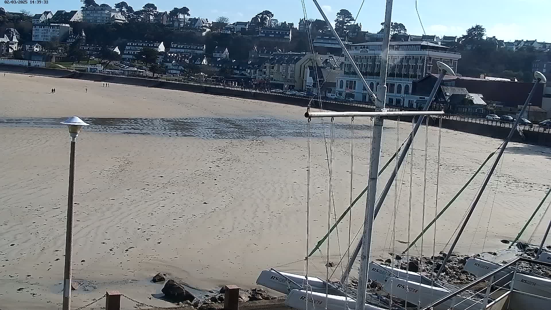 Several sailboats are grounded on a sandy beach at low tide, with a town and buildings visible in the background on a sunny day.