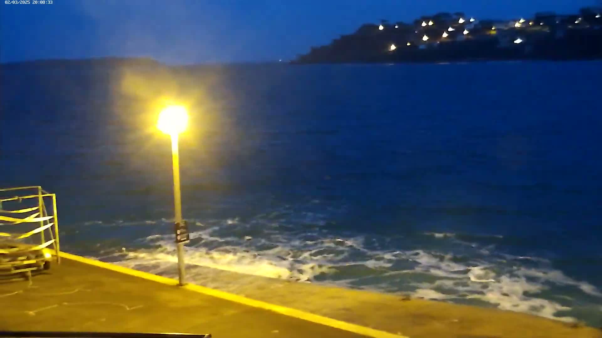 A nighttime view shows a dark body of water with small waves lapping against a concrete wall, illuminated by a single lamppost, and a distant hillside with lights visible.