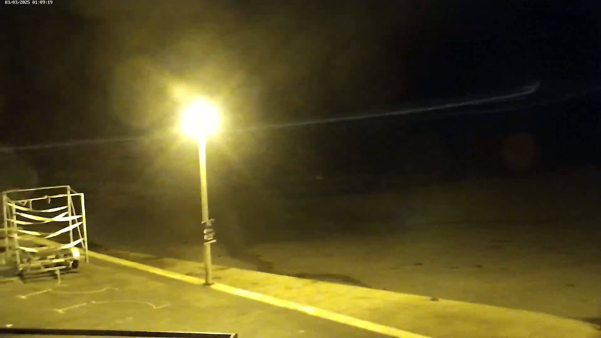 A nighttime outdoor scene shows a single lamppost illuminating an empty paved area, a metal frame structure to the left, and a hazy atmosphere.