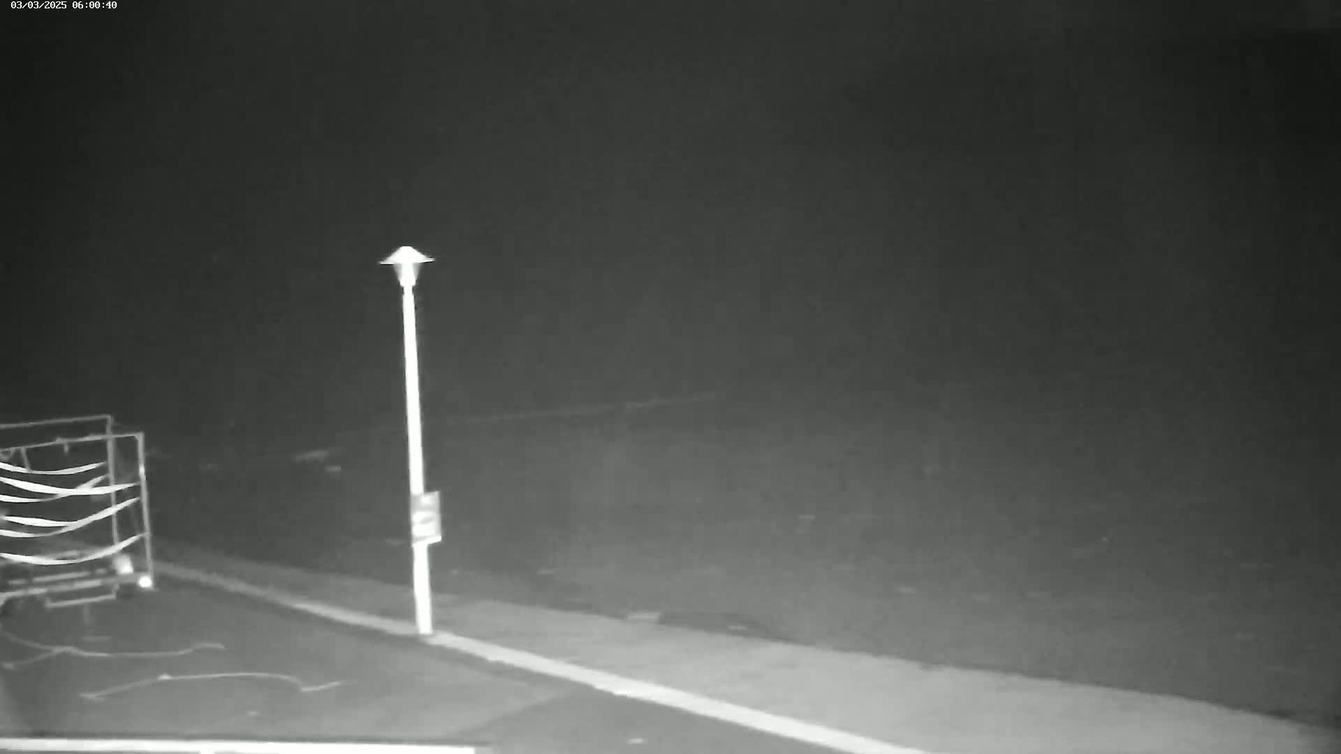 A nighttime, low-light image shows a tall lamp post next to a cart or small trailer in a mostly empty parking lot.