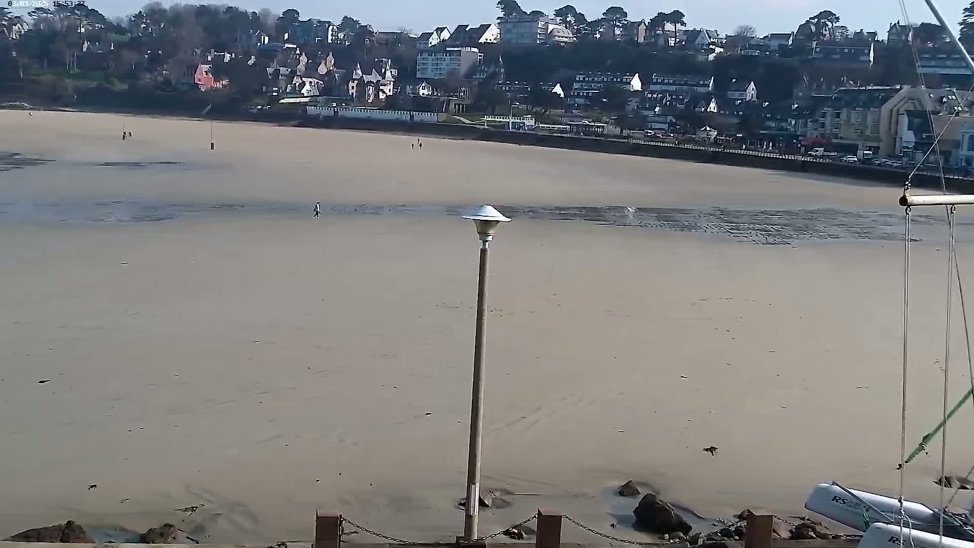 A mostly empty sandy beach stretches out from a shoreline of buildings under a sunny sky, with a tall lamppost in the foreground.