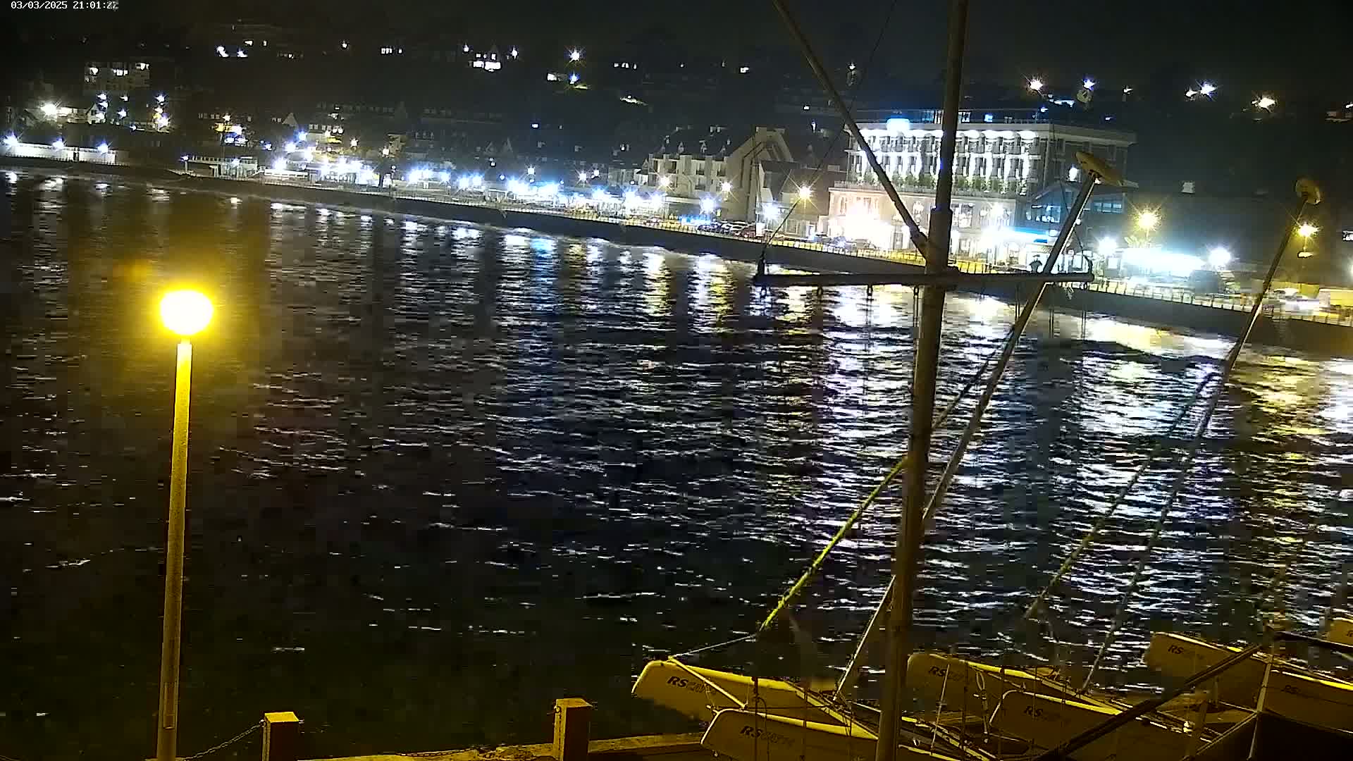 A nighttime view of a choppy waterfront with docked sailboats, illuminated buildings, and a single lamppost.