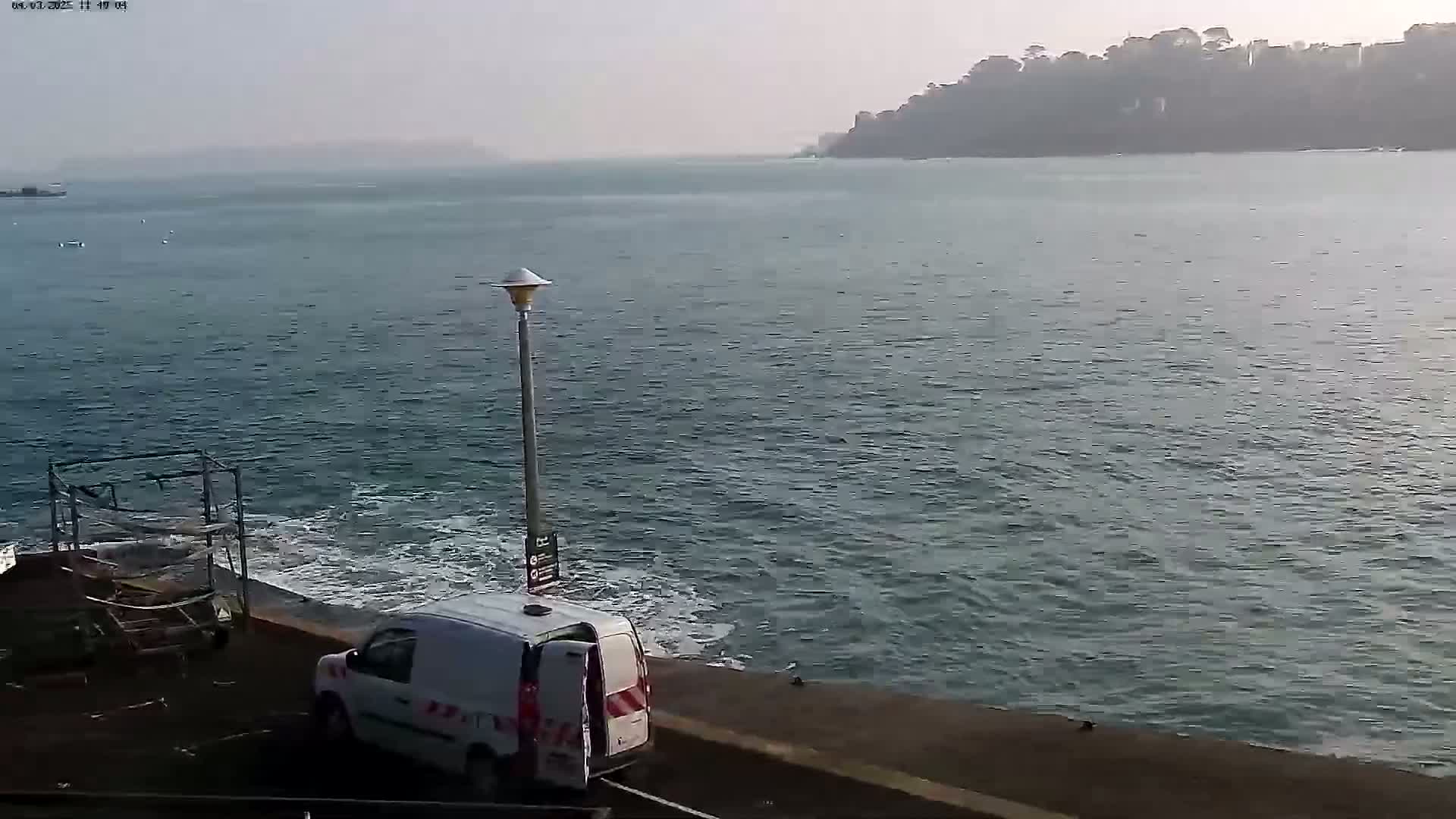 A white van is parked near a body of water with small waves lapping against a seawall, under an overcast sky, with a distant shoreline visible.