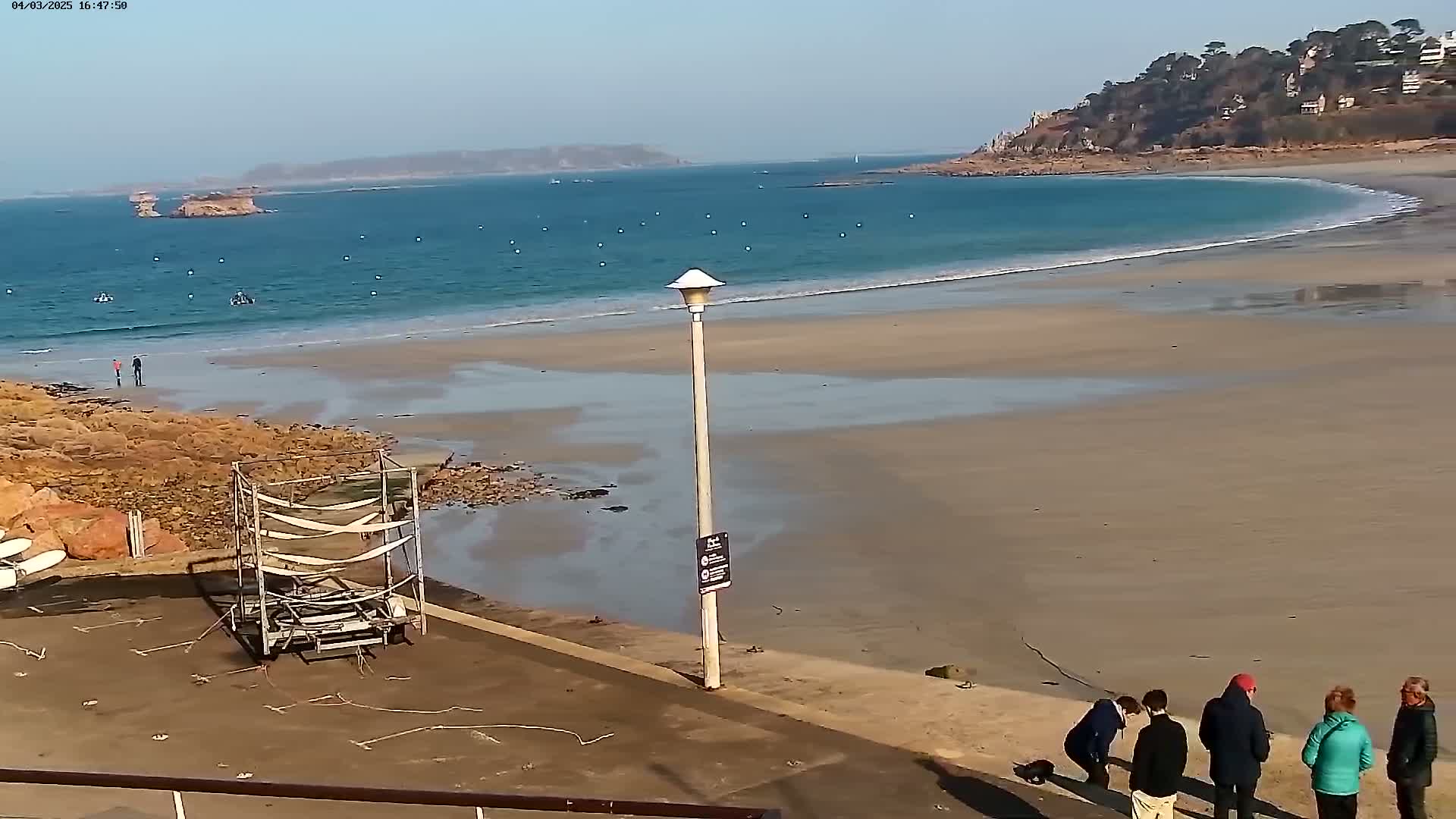 A sandy beach stretches along a calm ocean under a clear sky, with a few people standing near the water's edge and a lamppost nearby.