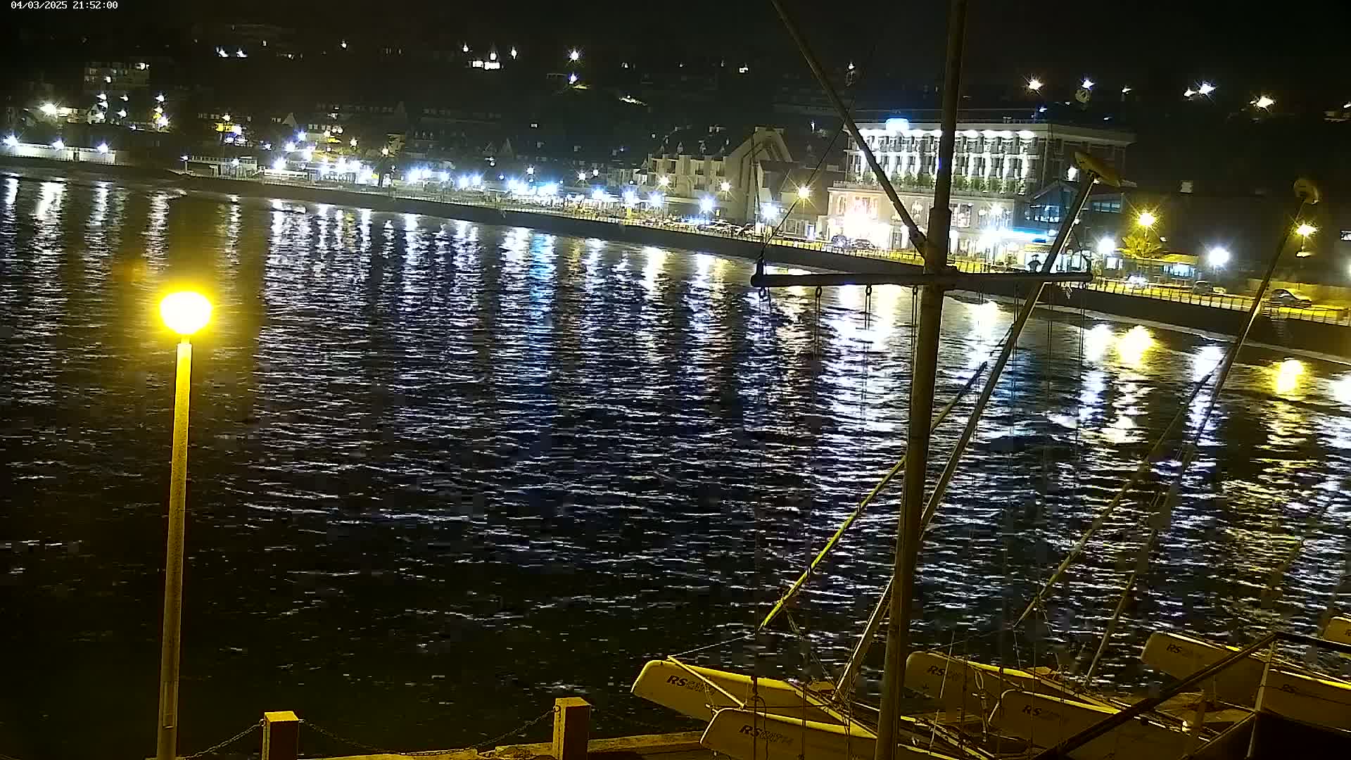 A nighttime view of a calm, dark body of water reflecting the lights of a seaside town, with several small sailboats moored at a dock in the foreground.