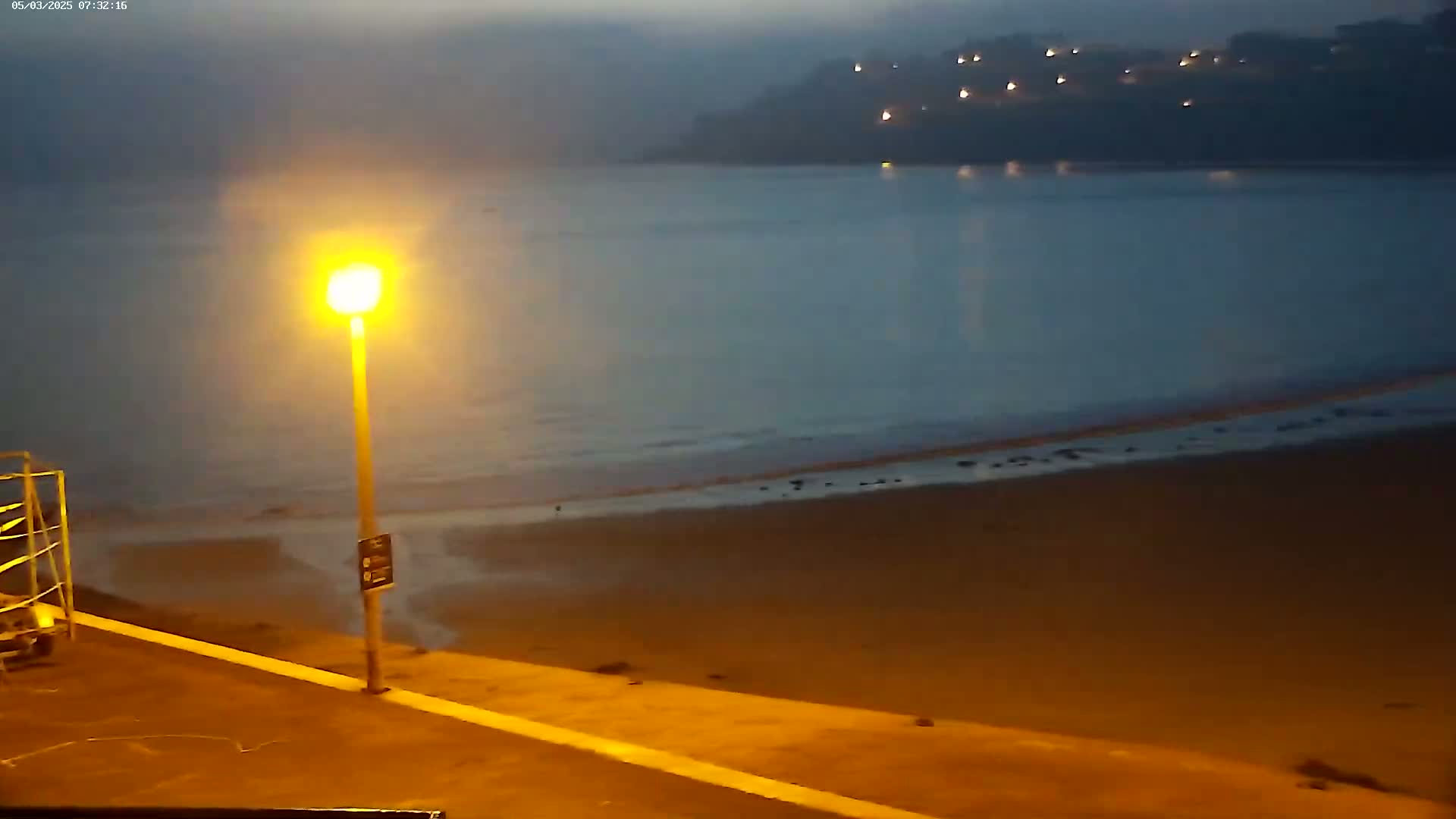 A nighttime view of a calm ocean beach under a dusky sky, illuminated by a single lamppost.