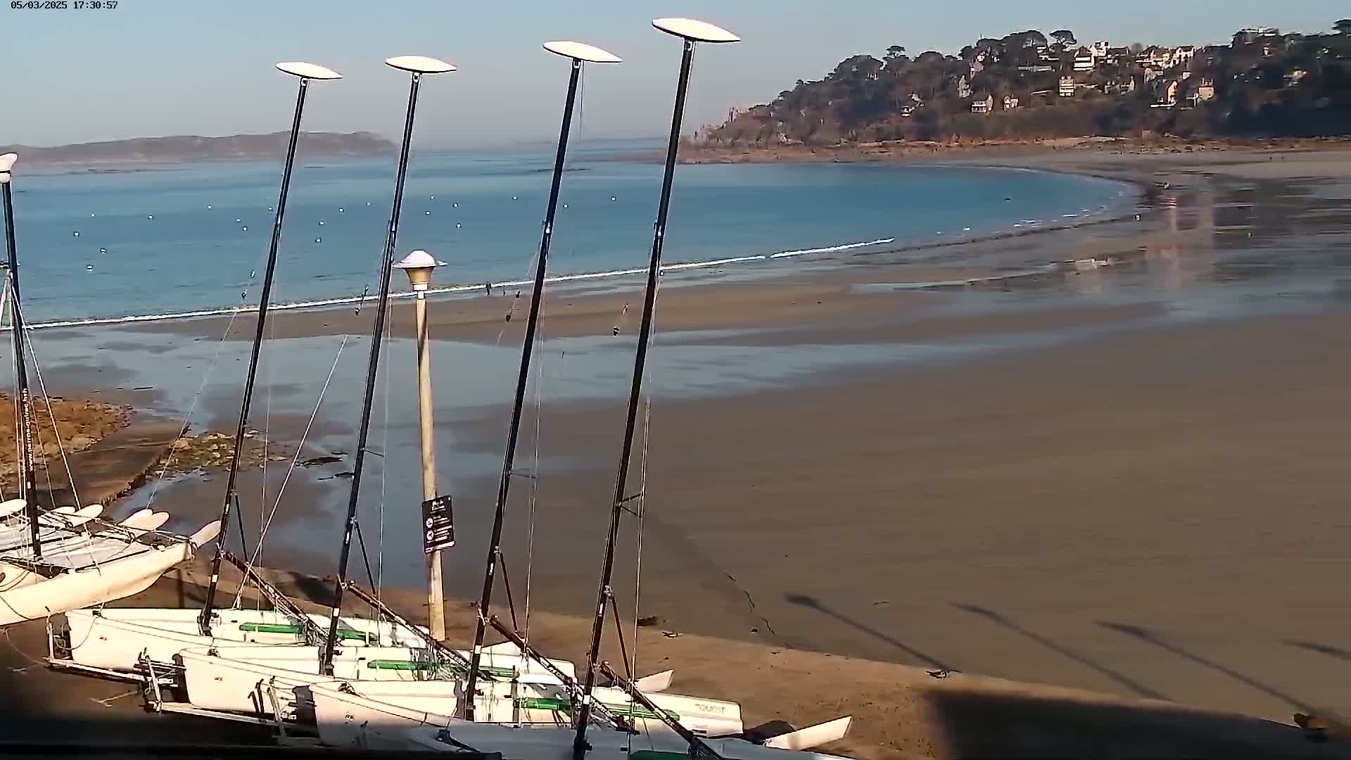 Several sailboats are beached on a sandy shore next to calm, blue water under a clear, sunny sky.