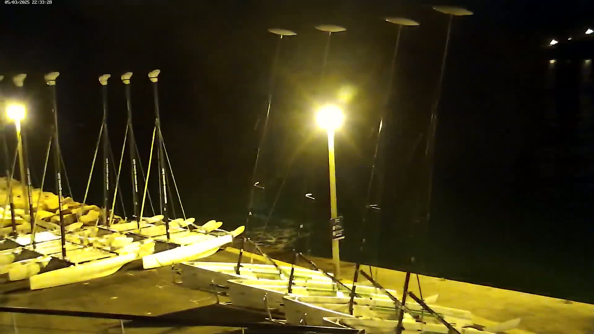 At night, numerous small sailboats are docked at a pier illuminated by bright lights.
