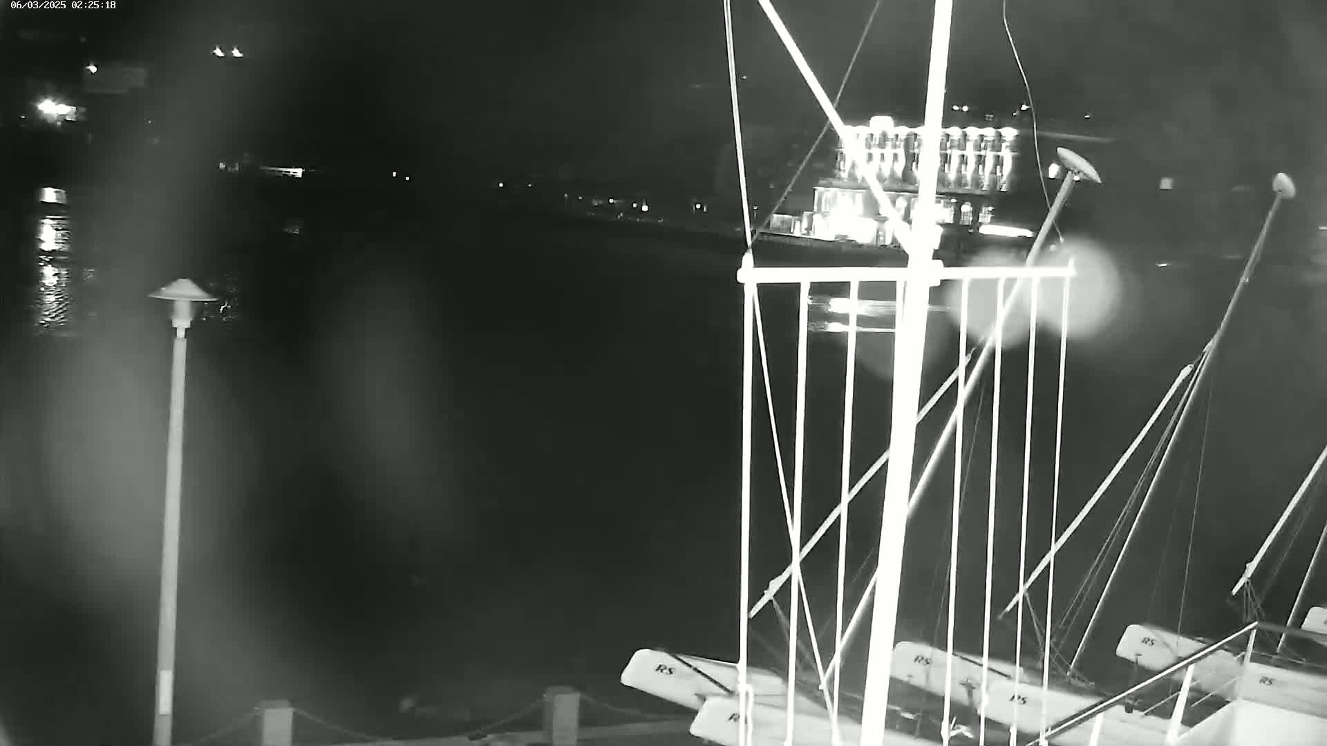 A nighttime, low-light view of several sailboats moored at a dock, with a blurry shoreline and water in the background.