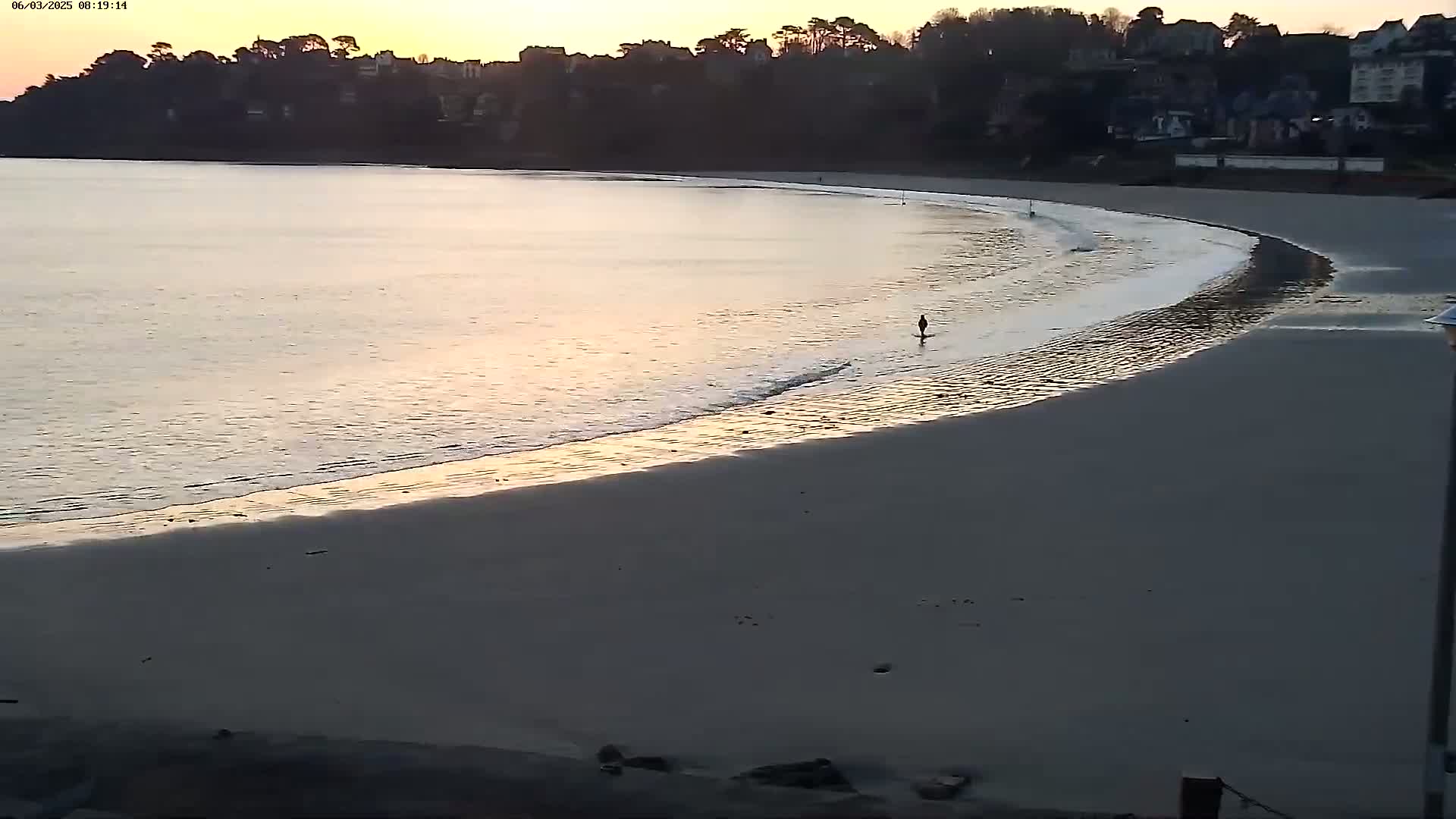 A person walks along a sandy beach curving around calm, pale-golden water at sunrise, with a tree-lined hillside visible in the background.