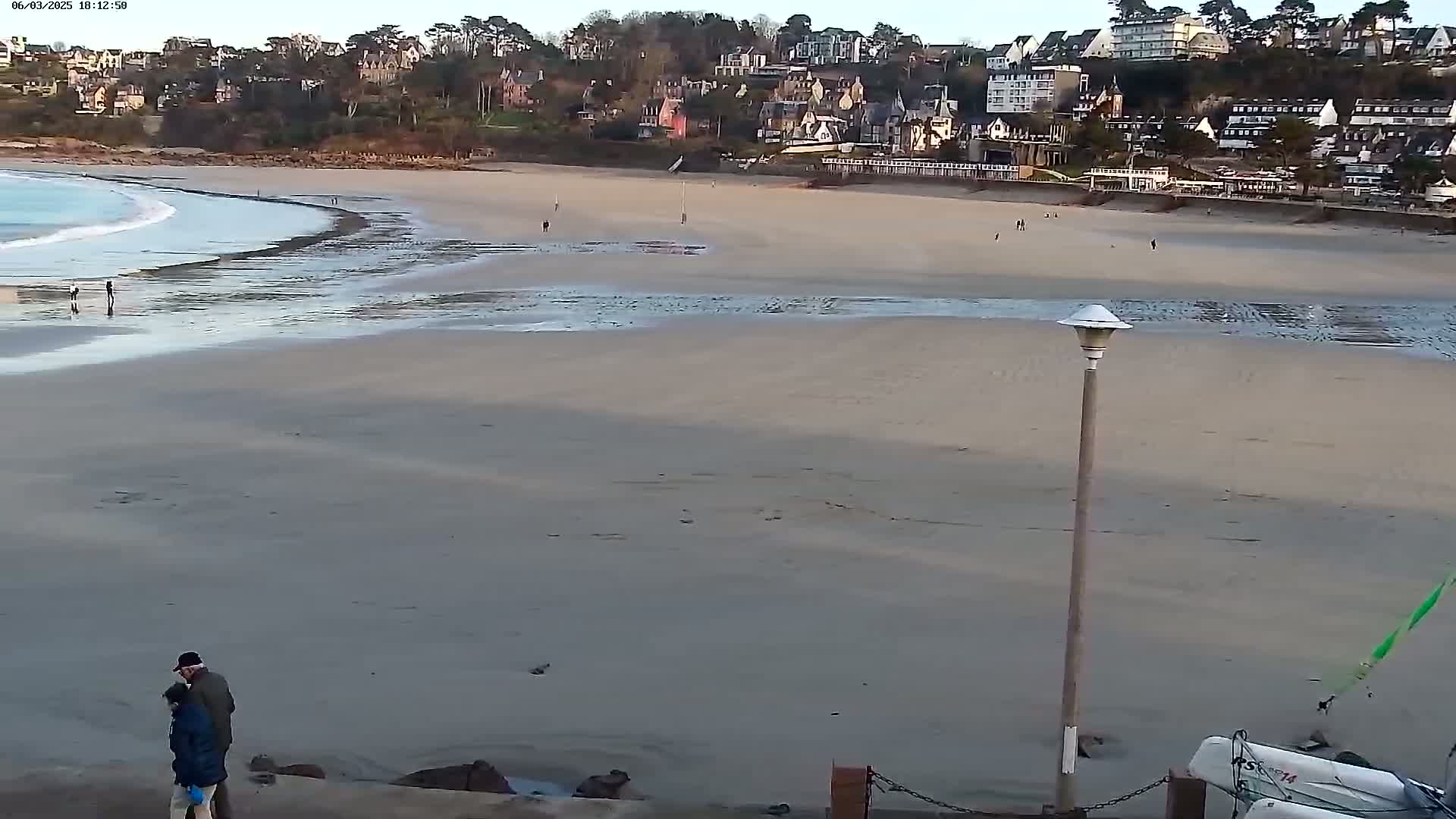 A wide shot of a sandy beach with calm waves, a few people, and a town of buildings overlooking the shore on a partly cloudy day.