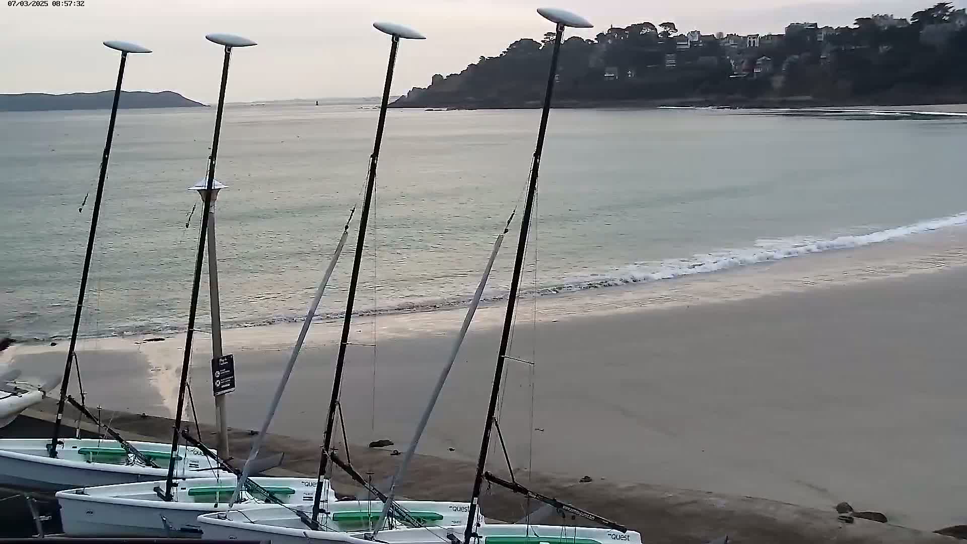 Several sailboats are beached on a sandy shore next to calm, light-grey water under an overcast sky.