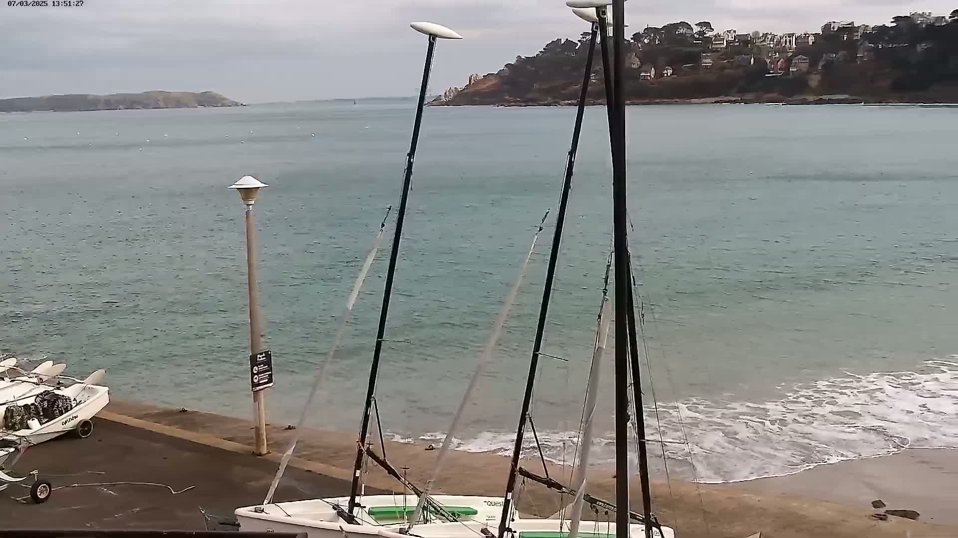 Two sailboats are beached on a sandy shore near a calm, light-green body of water under an overcast sky.