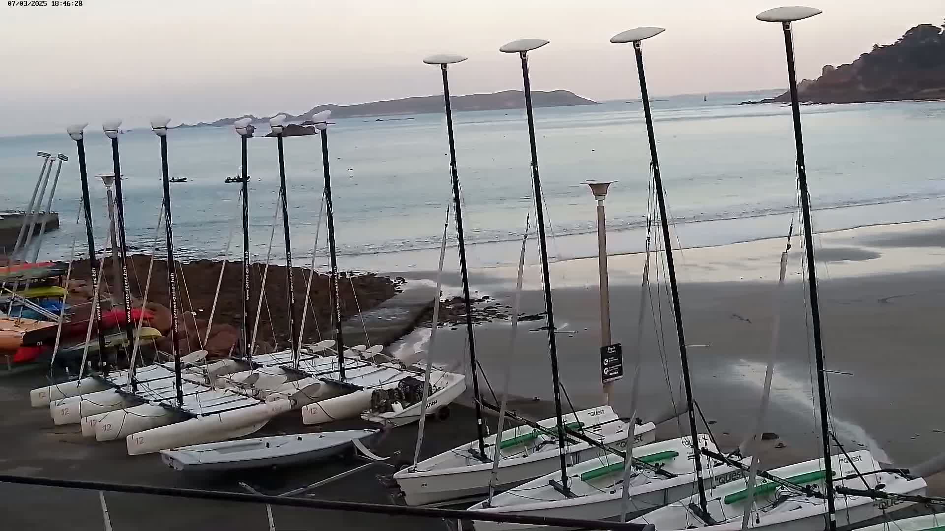 Numerous sailboats are beached on a sandy shore under a cloudy sky, with calm ocean waters visible in the background.