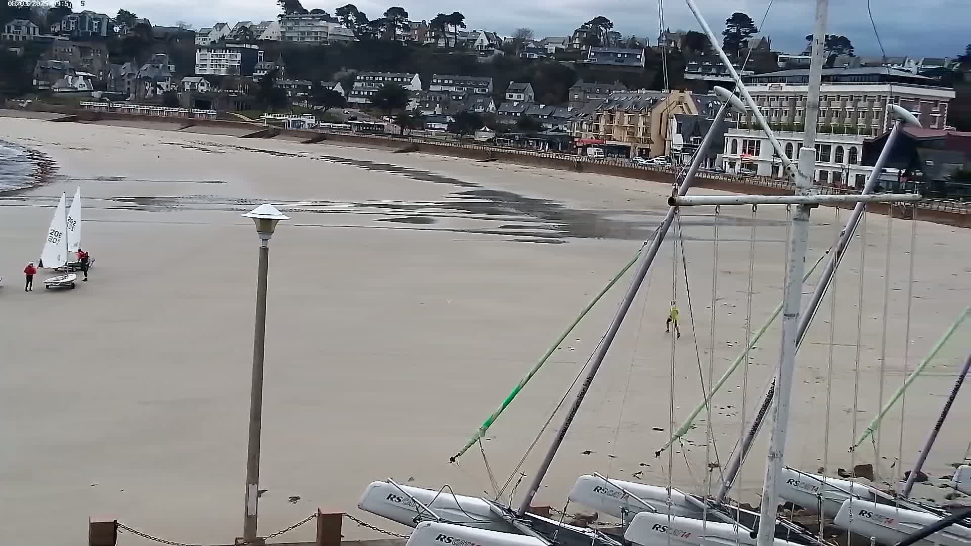 A sandy beach with several sailboats beached on the sand, a few people are near the boats and a town is visible in the background under an overcast sky.