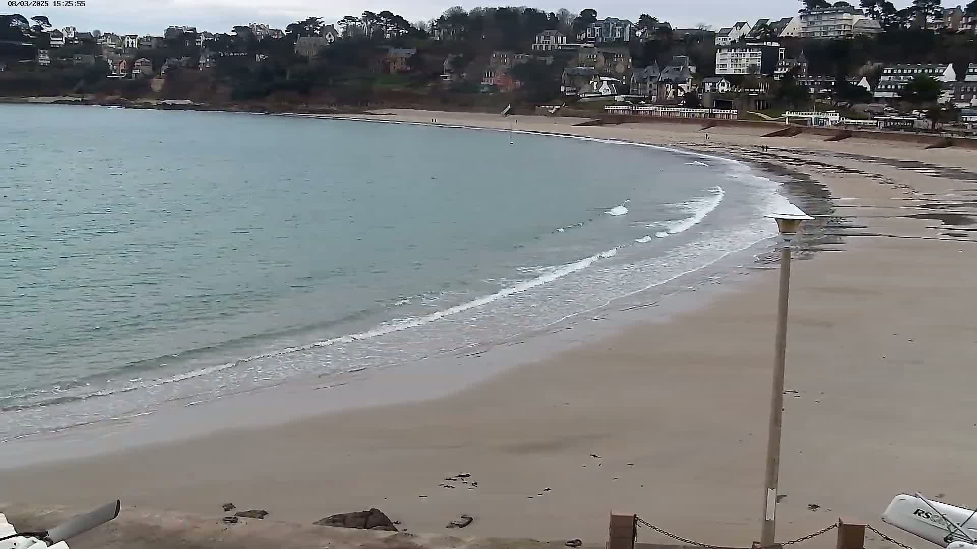 A sandy beach curves gently into a calm, light-blue sea under an overcast sky, with a line of houses visible on a hillside in the background.