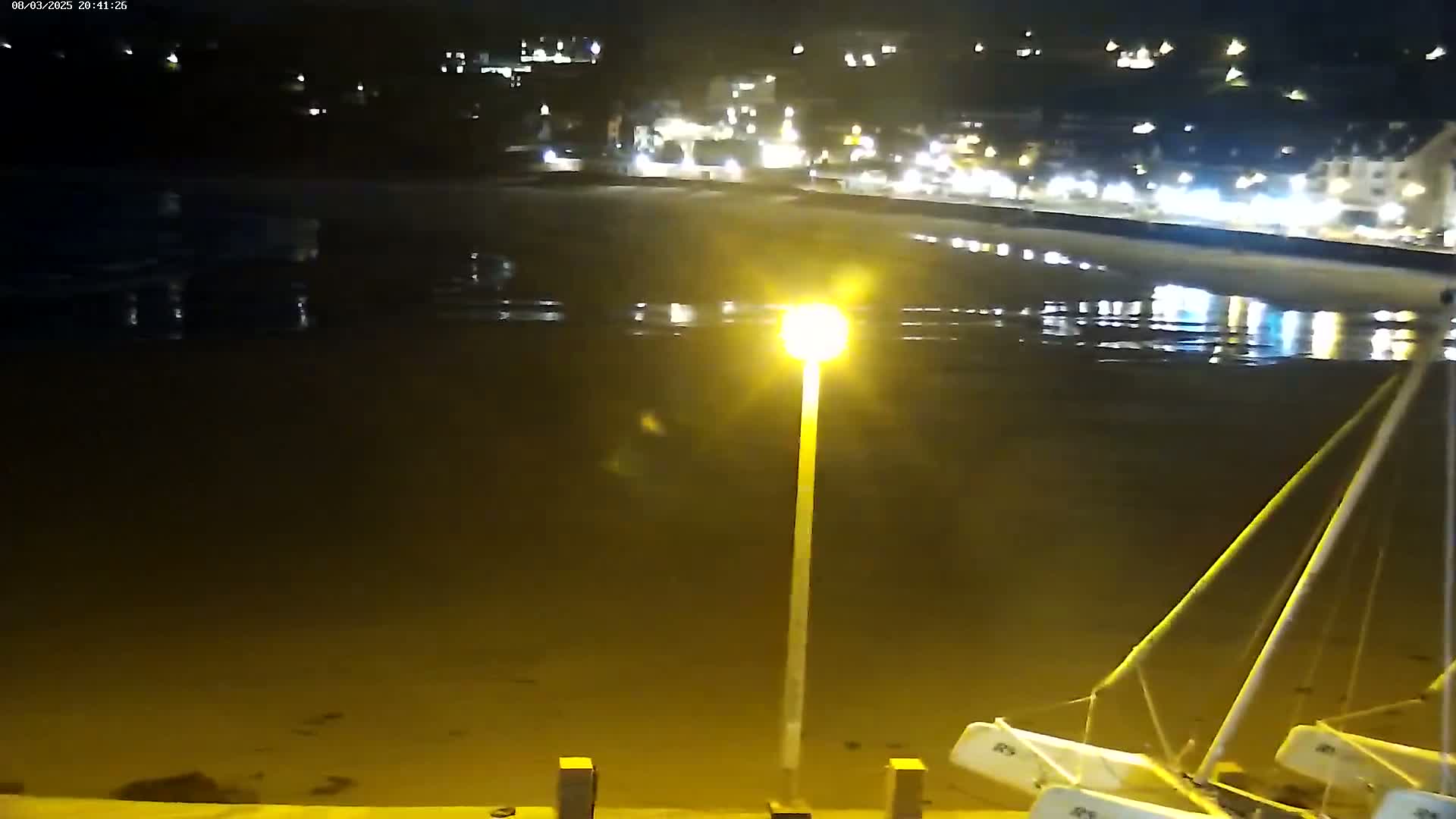 A nighttime view of a beach at low tide, with several small boats beached nearby and a town's lights reflected in the wet sand.