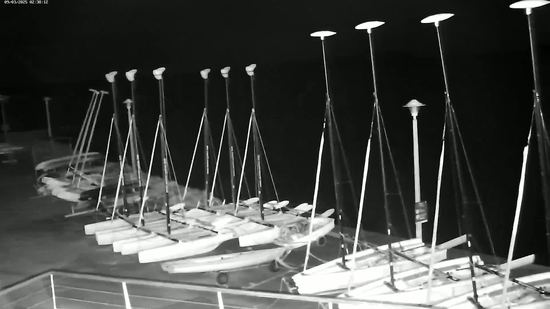 A nighttime, black and white image shows numerous small sailboats docked at a marina under several tall lights.
