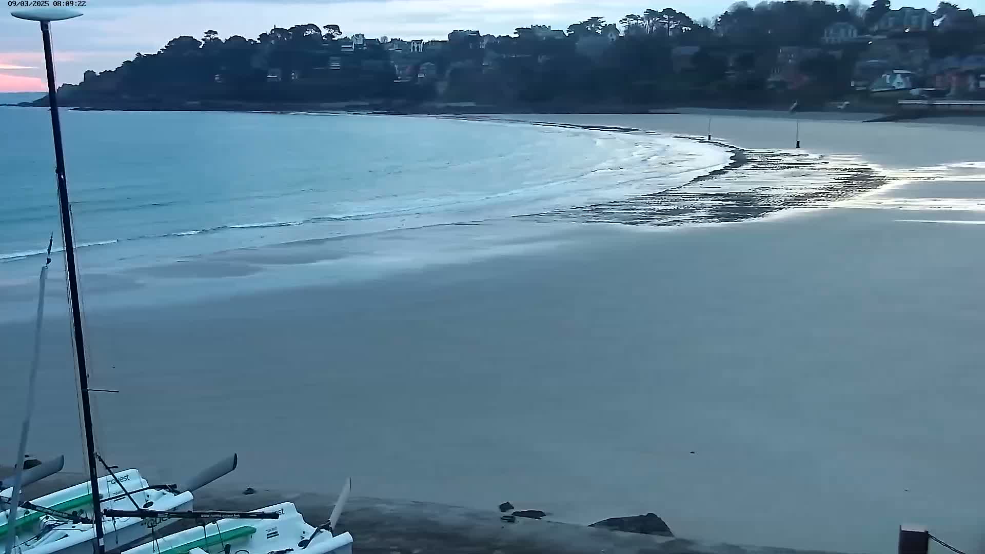 A long, sandy beach curves around a calm bay under an overcast sky, with two small sailboats beached nearby.