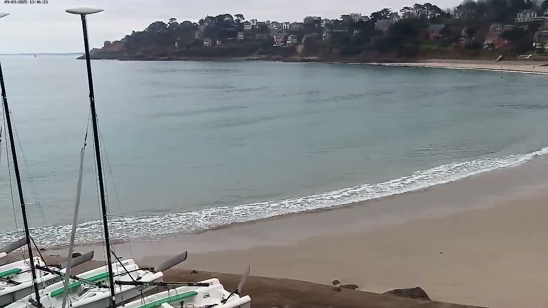 Several small sailboats are beached on a sandy shore next to calm, light-blue water lapping gently at the sand, with a residential area visible across the bay under an overcast sky.