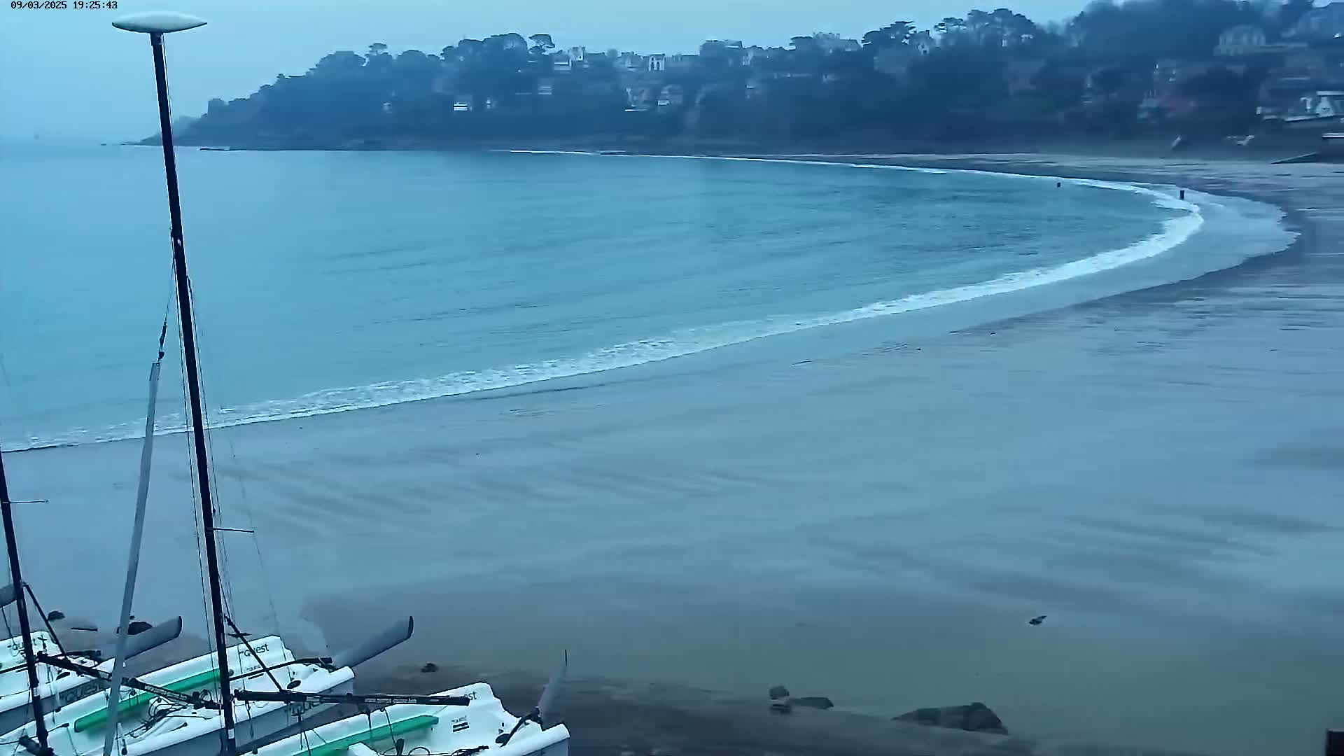 Several sailboats are beached on a sandy shore next to a calm, blue ocean under an overcast sky.
