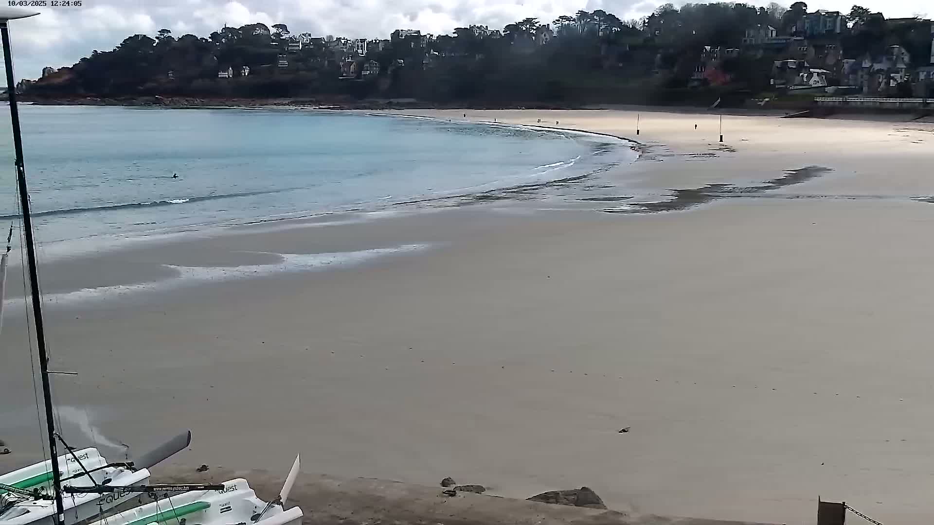 A sandy beach curves around a calm, light-blue bay under a mostly cloudy sky, with small waves lapping the shore and a few people visible in the distance.
