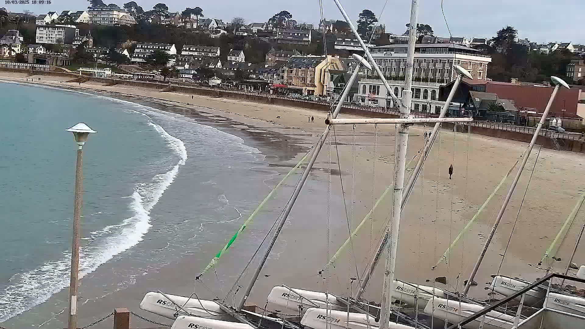 Several sailboats are beached on a sandy shore next to a calm sea with small waves, with a town visible on a hillside in the background under an overcast sky.
