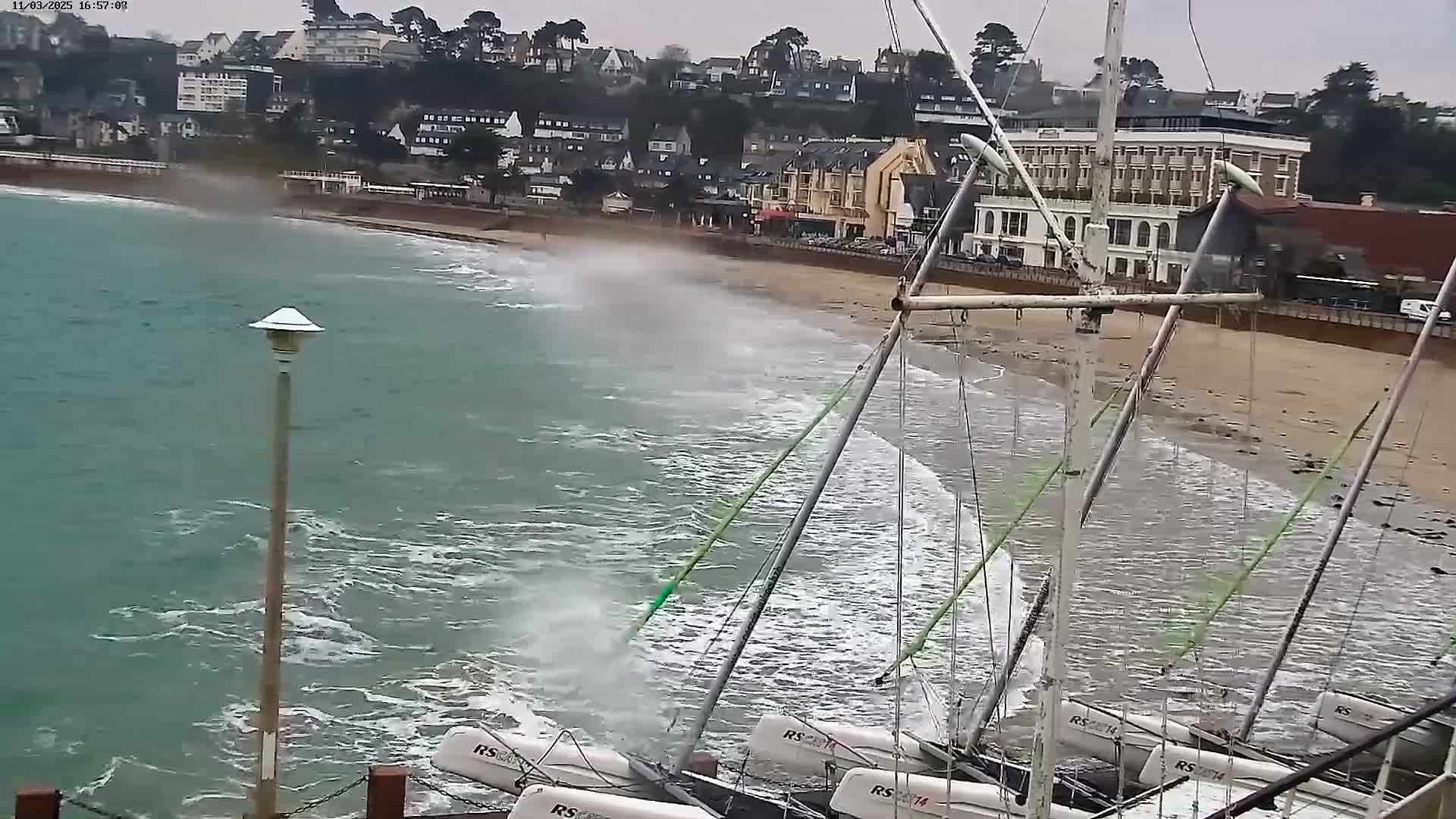 Several sailboats are beached on a sandy shore near a town under overcast skies with choppy waves crashing against the beach.
