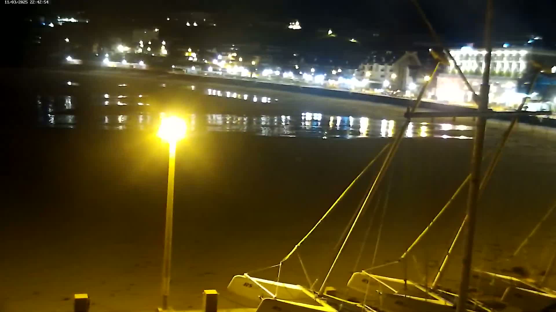 At night, several sailboats are beached on a sand-covered shore near a town with illuminated buildings and a calm, reflecting body of water.