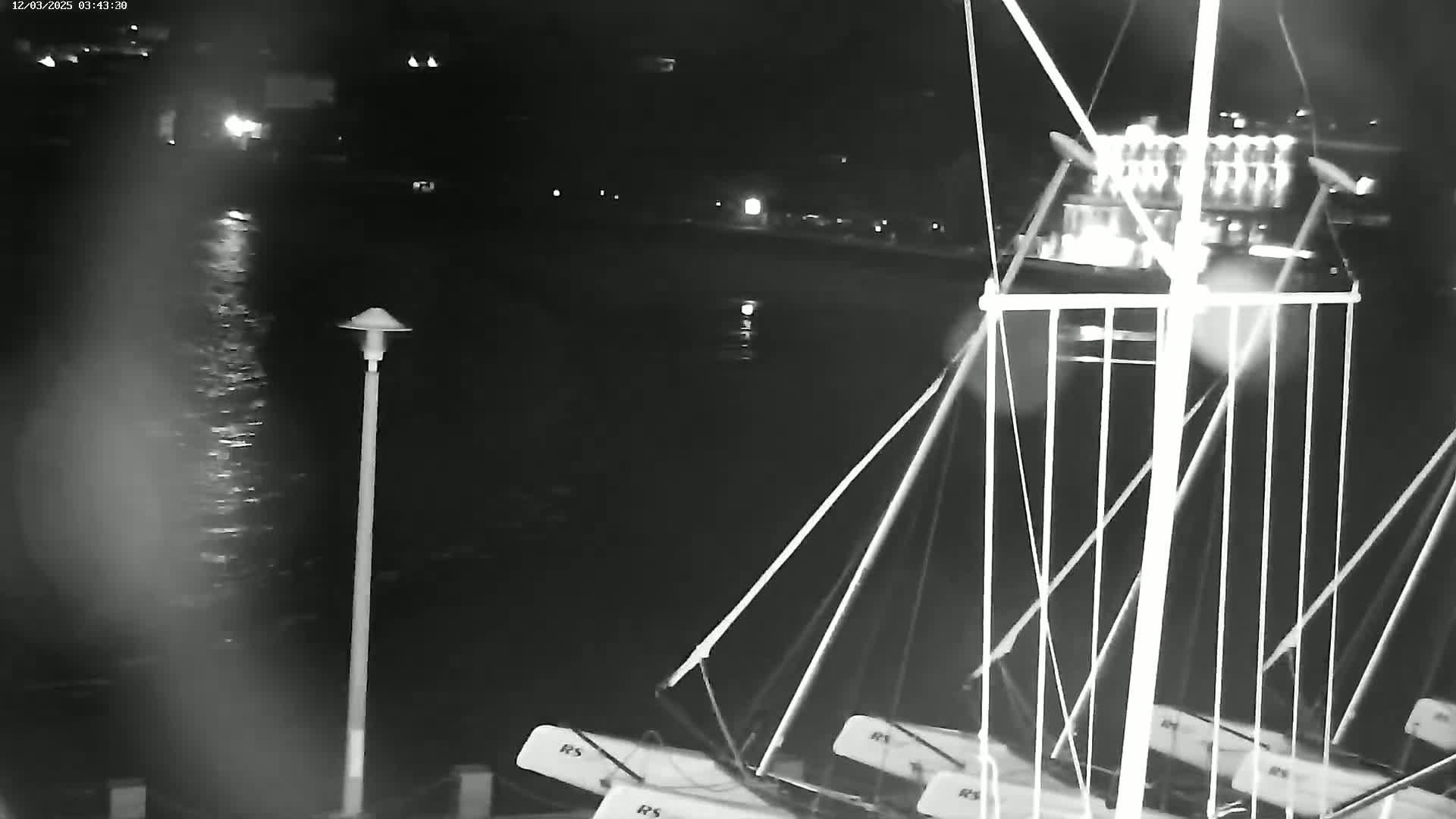 A nighttime, low-light image shows several small sailboats moored at a dock next to a body of dark water, with a single lamppost visible on the shore.