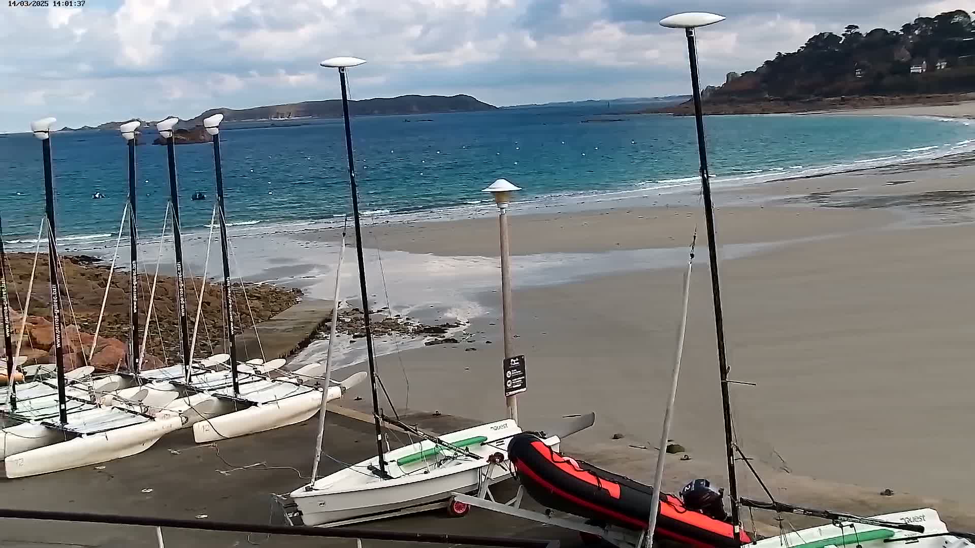 Several small sailboats are beached on a sandy shore next to a calm, blue ocean under a partly cloudy sky.
