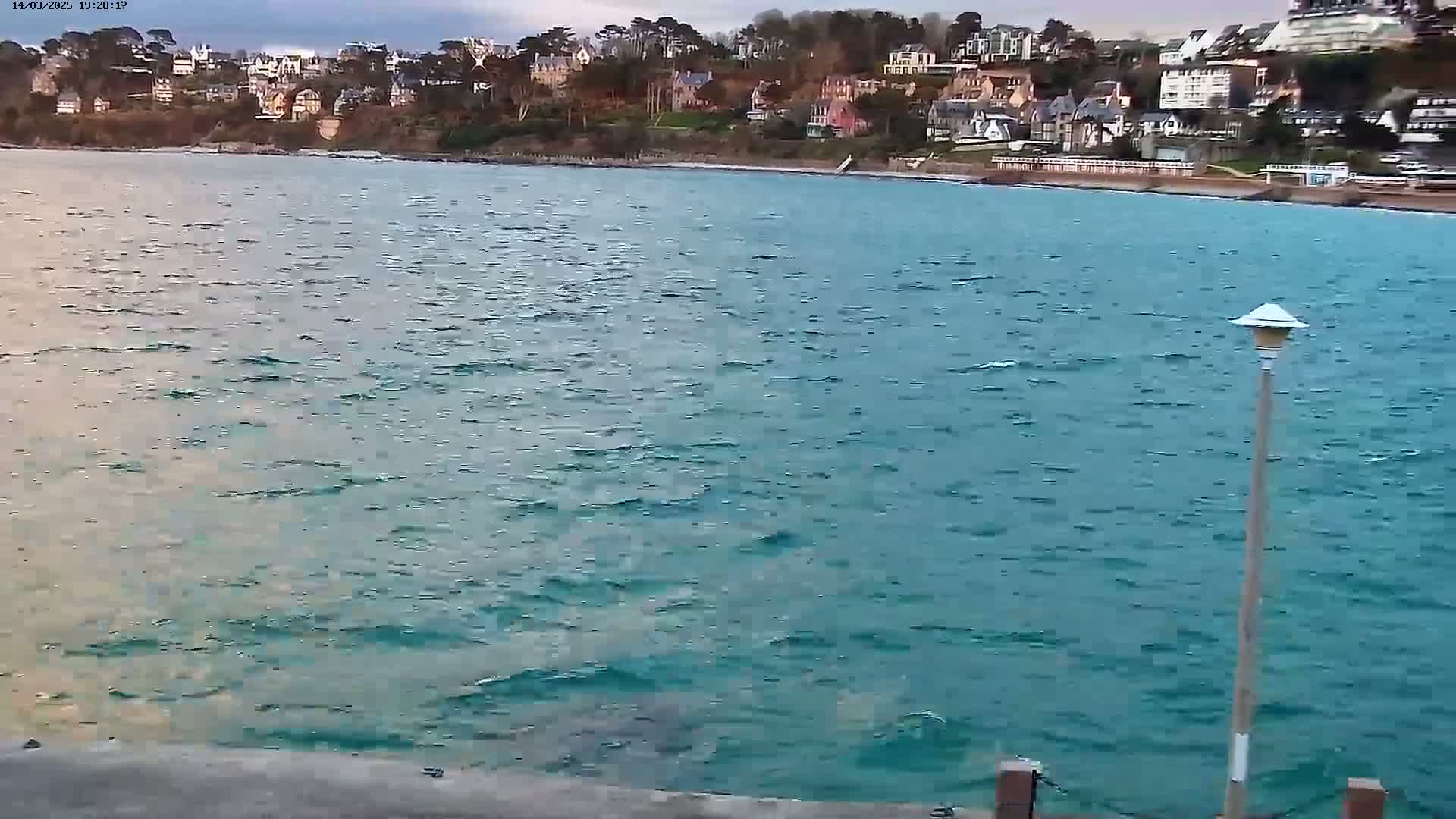 A body of choppy teal water stretches out before a line of buildings nestled on a hillside.