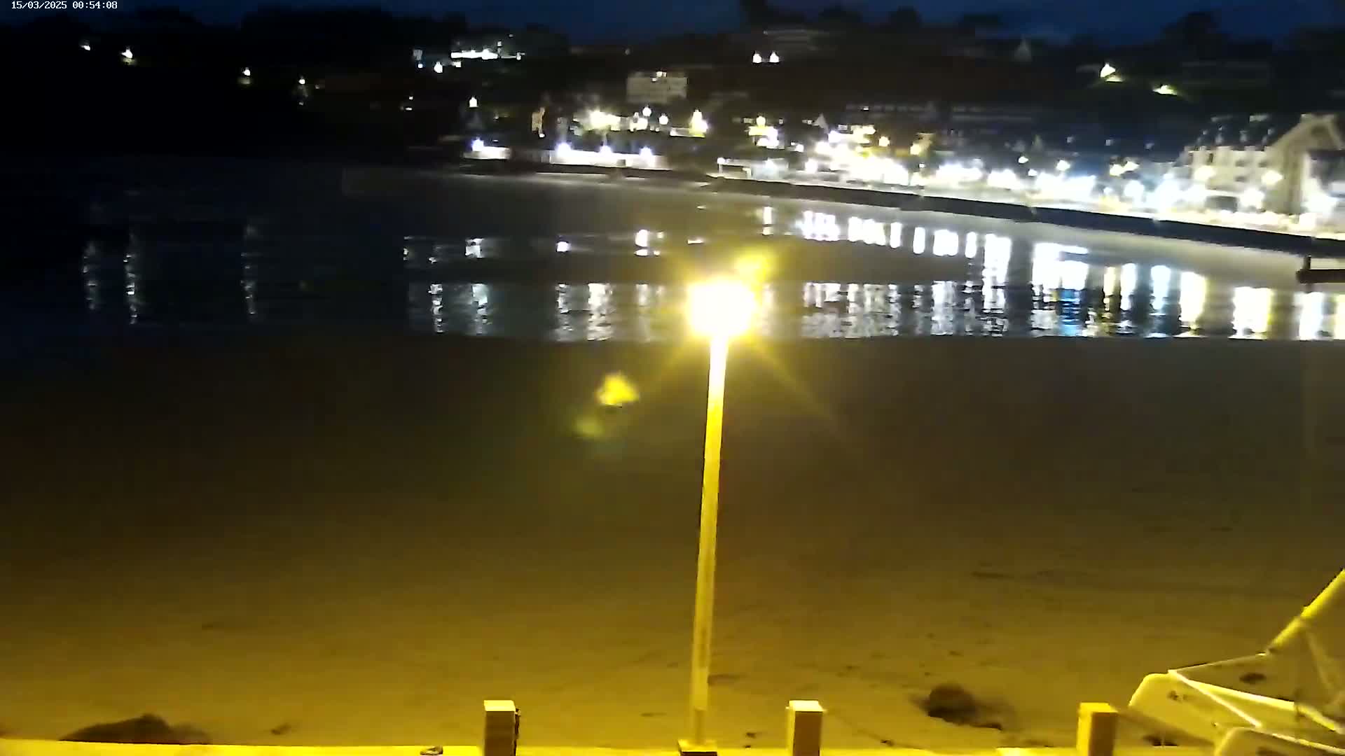 A nighttime view of a calm body of water reflecting the lights of a town on the opposite shore, illuminated by a single lamppost on a sandy beach.