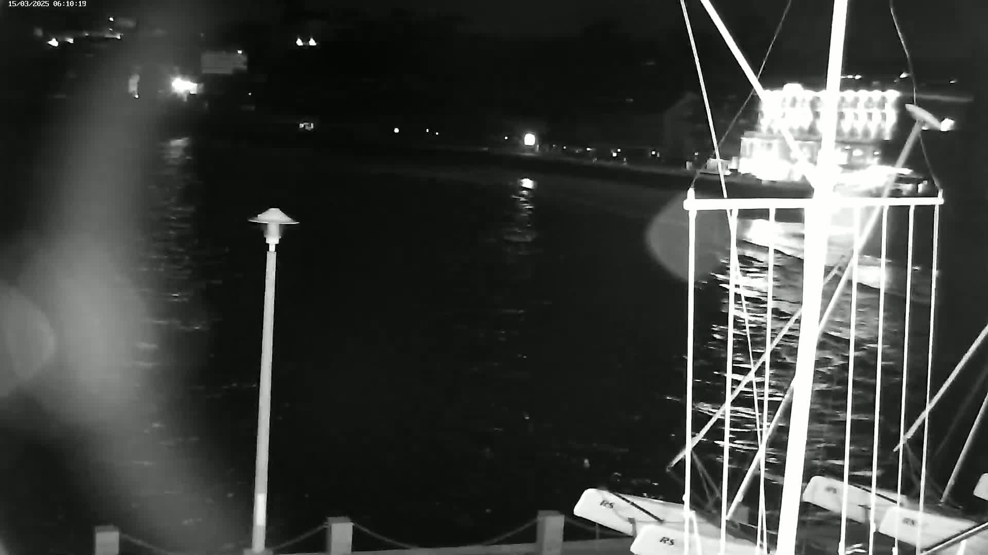 A nighttime, low-light view shows a body of water with a boat and a lit building in the background, along with a pier and a lamppost.