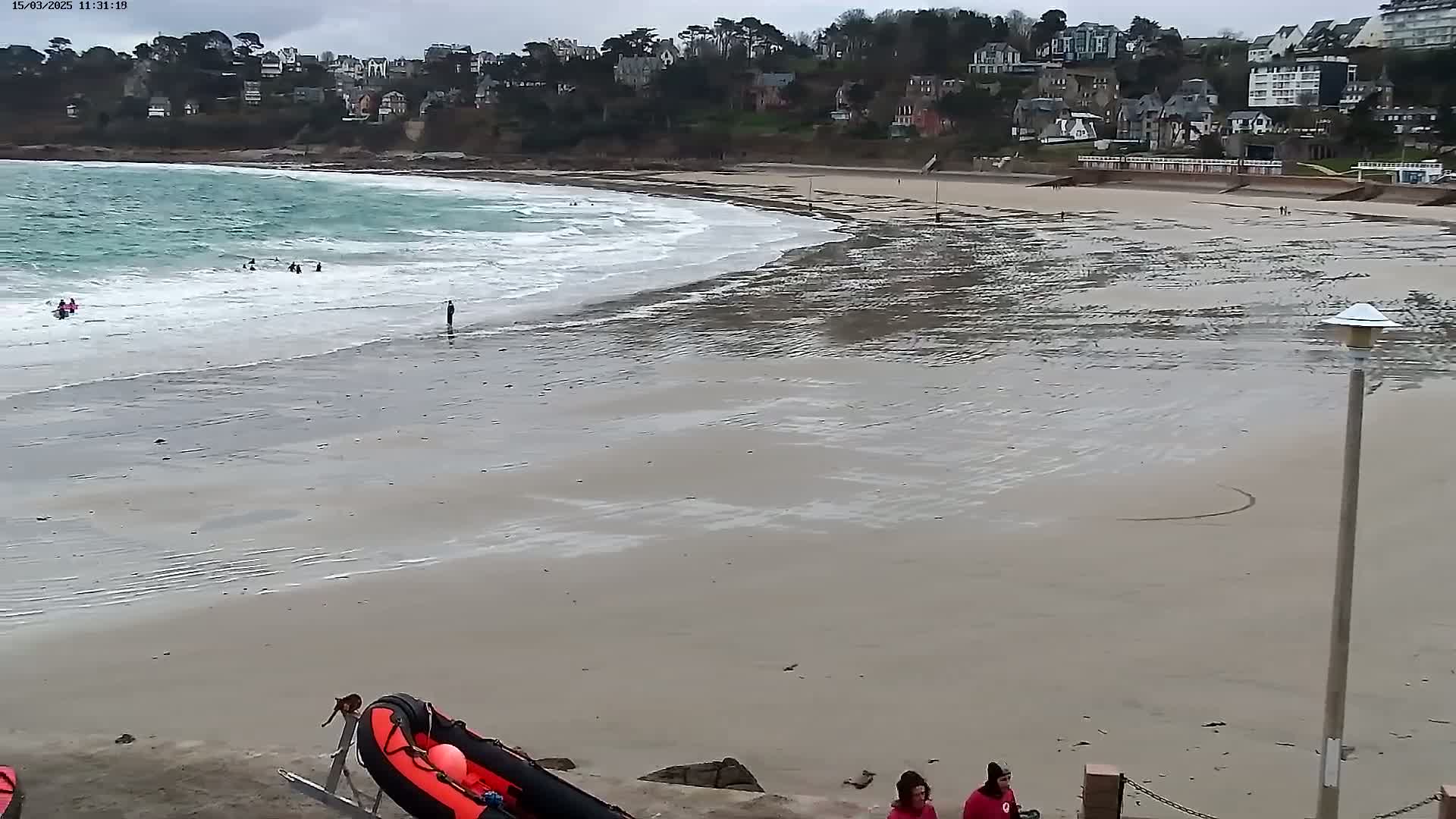 A sandy beach with small waves under an overcast sky, showing several people in the water and on the shore near a red rescue boat.