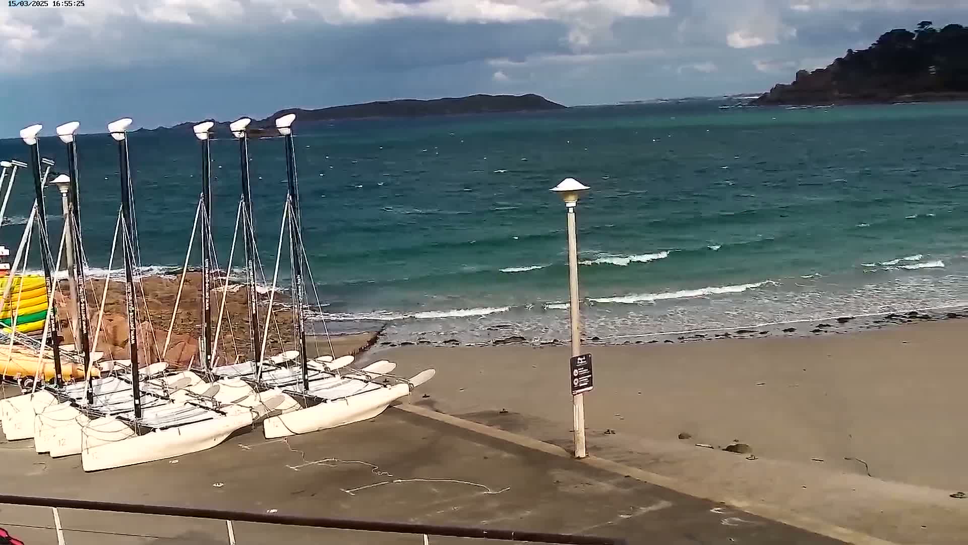 Several catamarans are docked on a sandy beach next to a body of water with small waves under a partly cloudy sky.