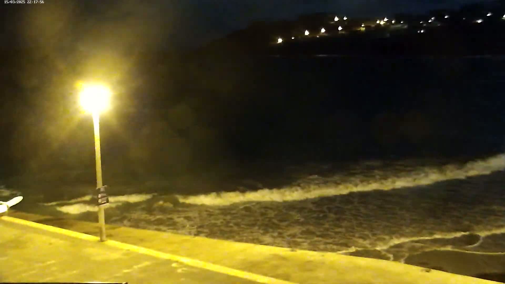 A nighttime view of moderate waves crashing on a shoreline near a bright streetlight, with distant lights visible on a hilltop.